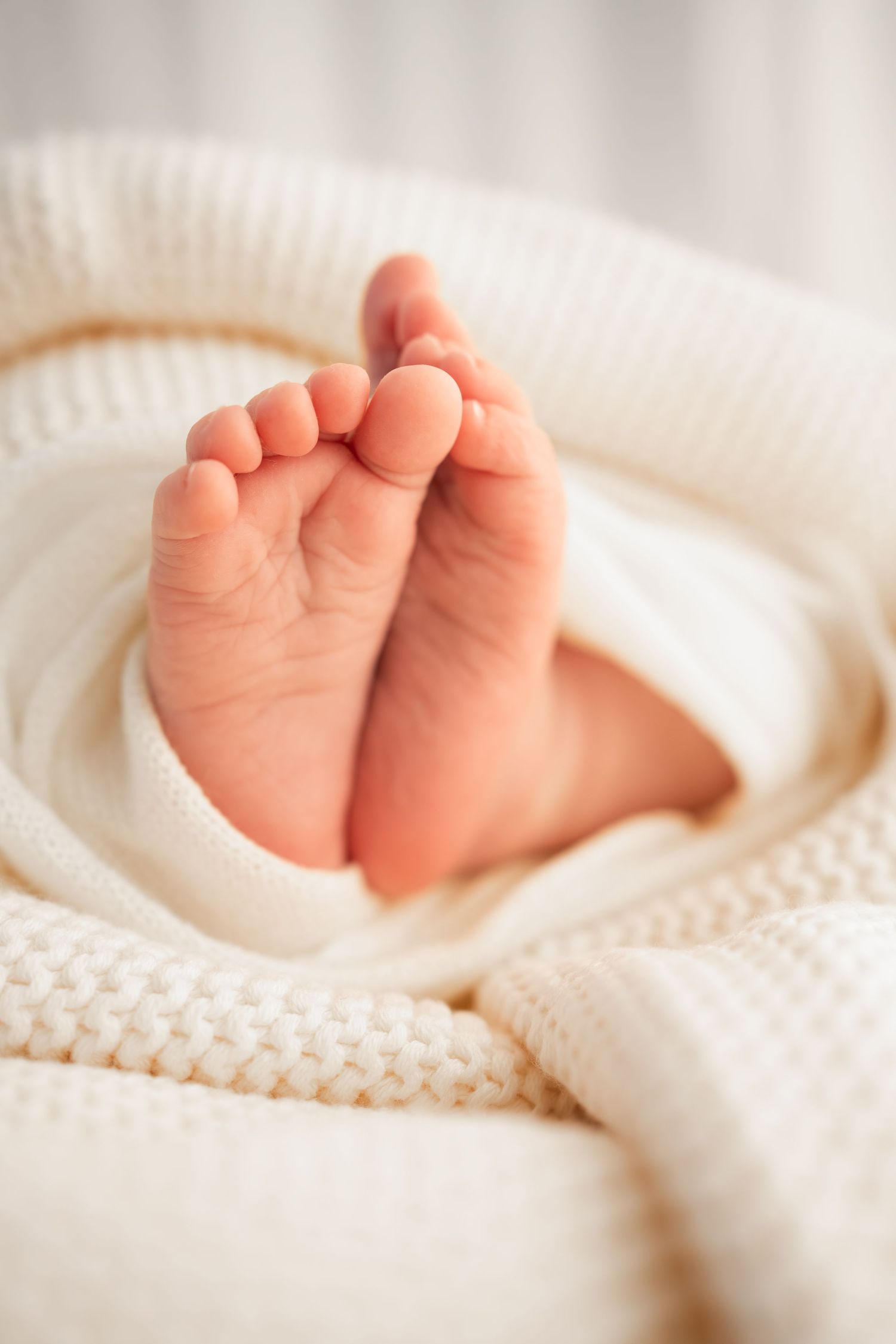 Close-up of tiny newborn baby feet peeking out from a soft cream knit blanket, with a blurred neutral background.