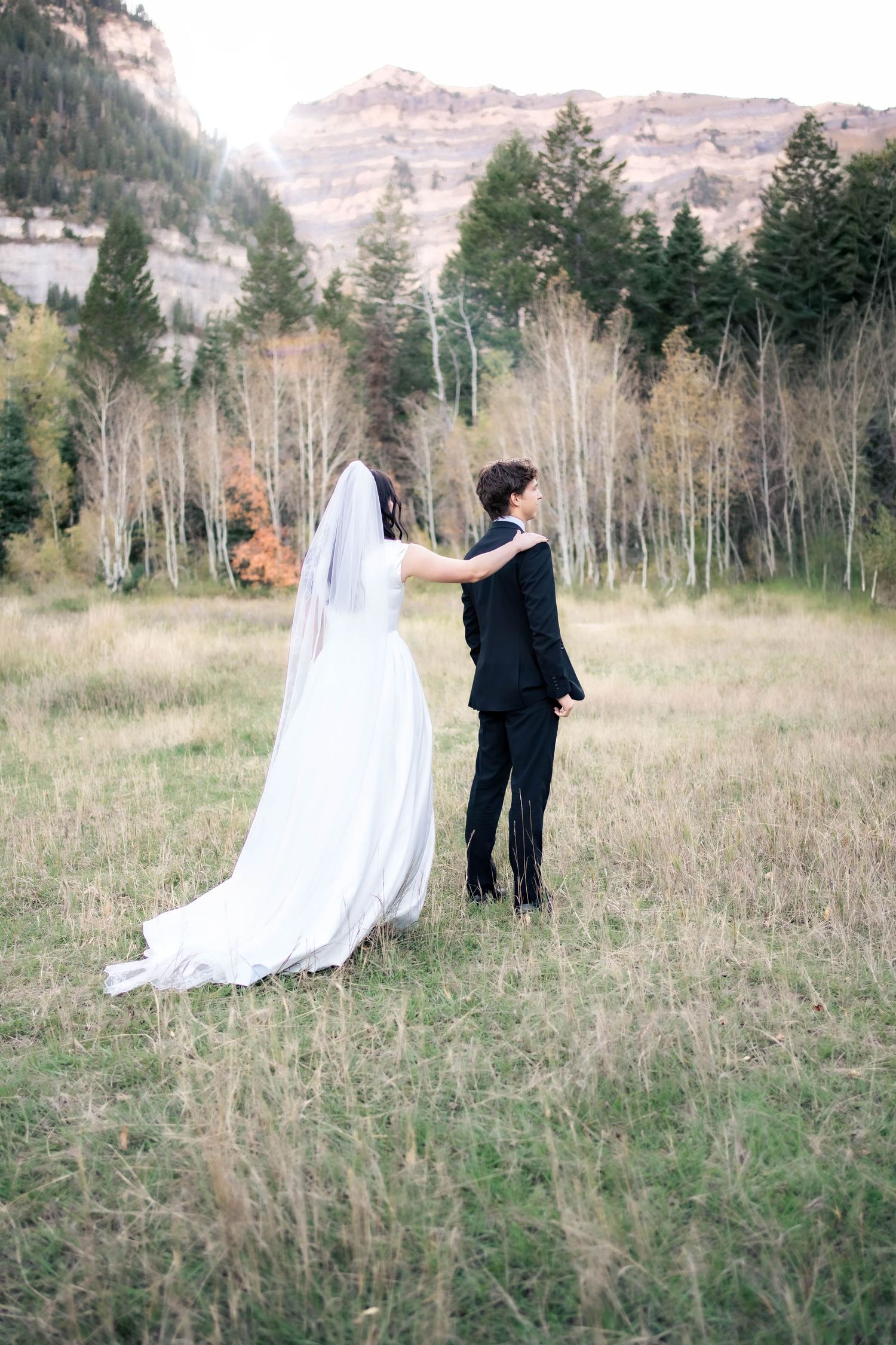 Bride in a flowing white gown and long veil reaches out to a groom in a dark suit in a grassy mountain meadow with autumn aspen trees.