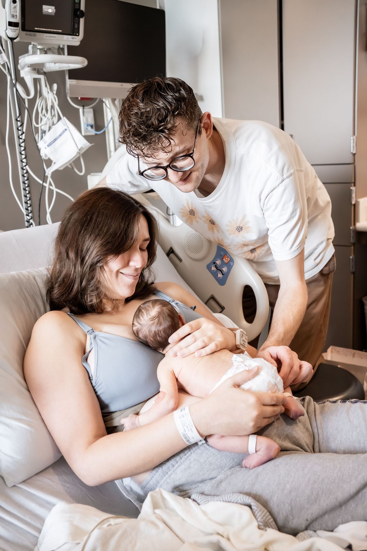 New parents smile at their newborn baby in a hospital room, mother resting in bed while father leans in close.