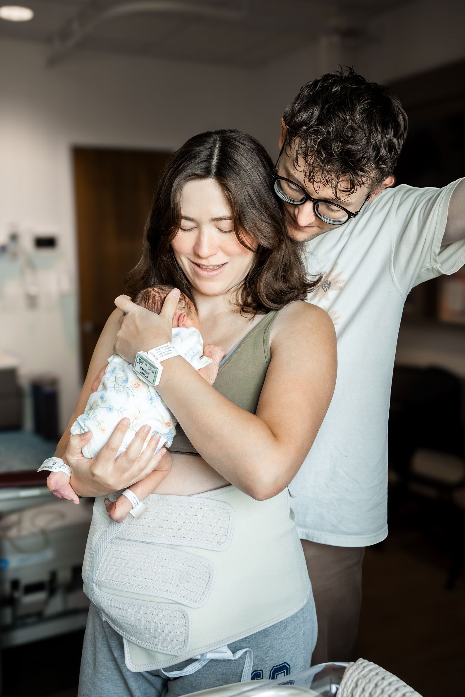 New parents in hospital room, mother holding newborn baby while father lovingly looks over her shoulder.