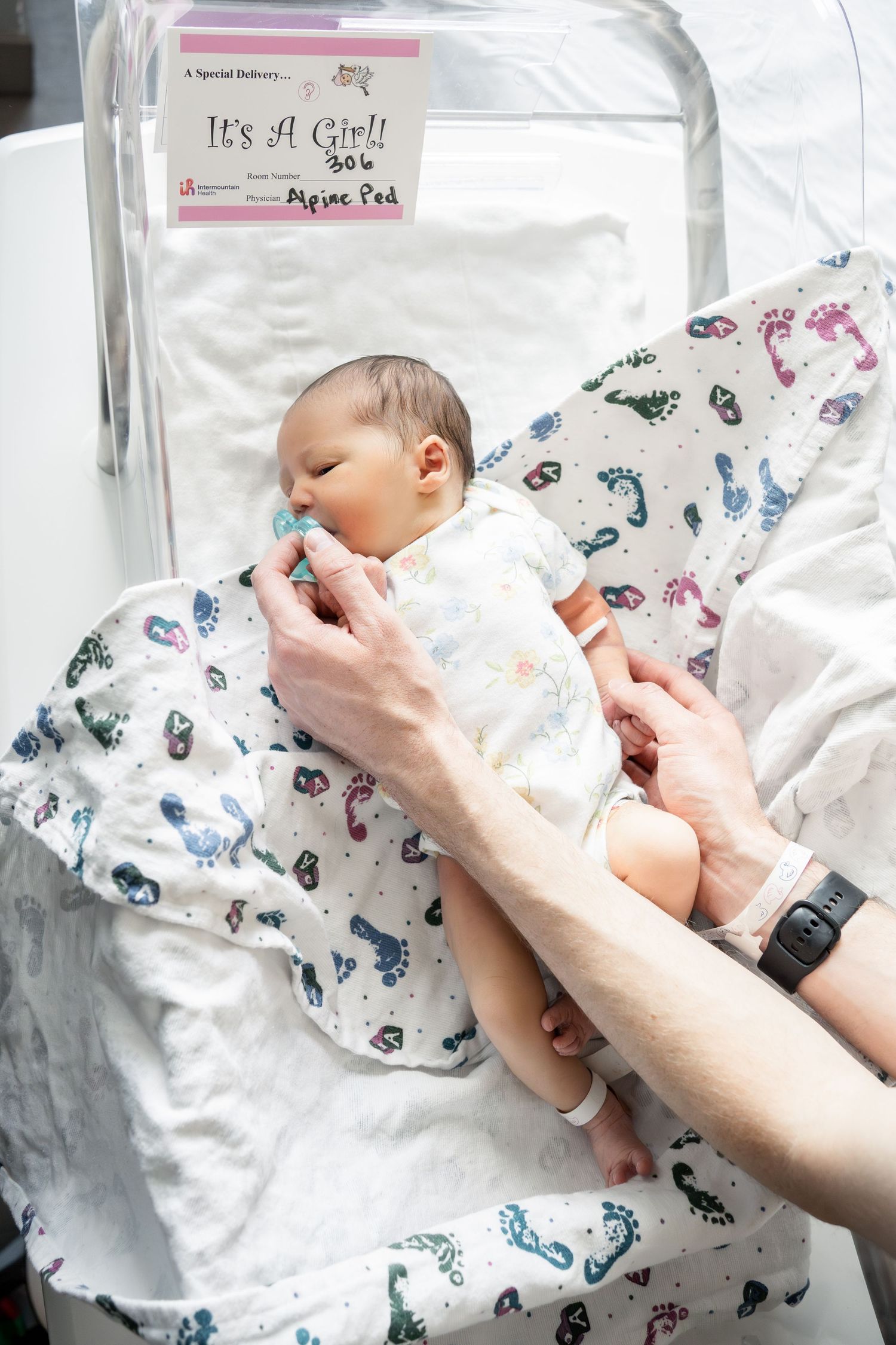Newborn baby girl wrapped in a floral swaddle blanket in a hospital bassinet with an It's A Girl sign.