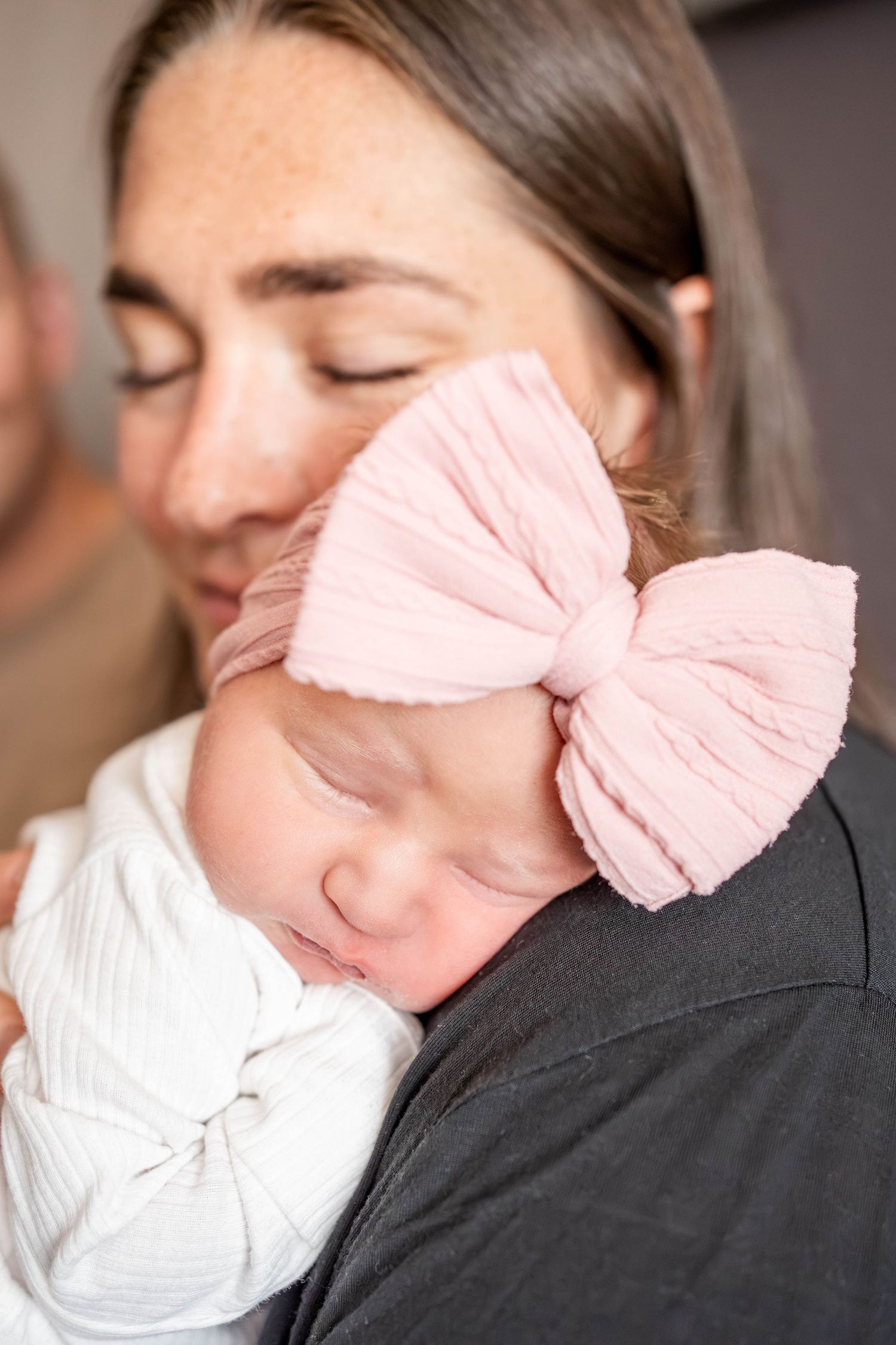 A mother with closed eyes tenderly holds her sleeping newborn baby wearing a white swaddle and pink bow headband.