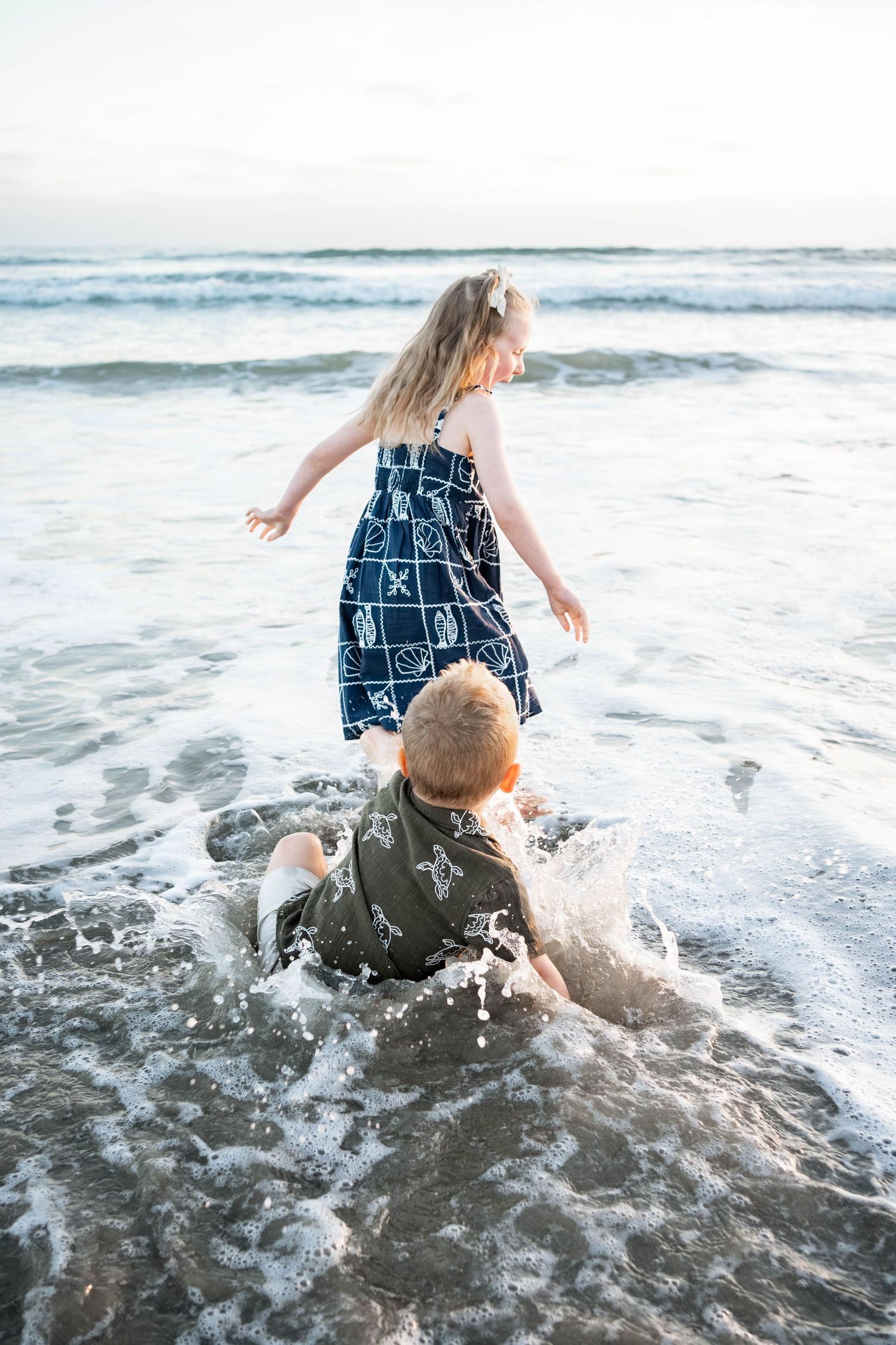 Two children playing in ocean waves on a beach, girl in blue dress and boy sitting in the surf splashing.