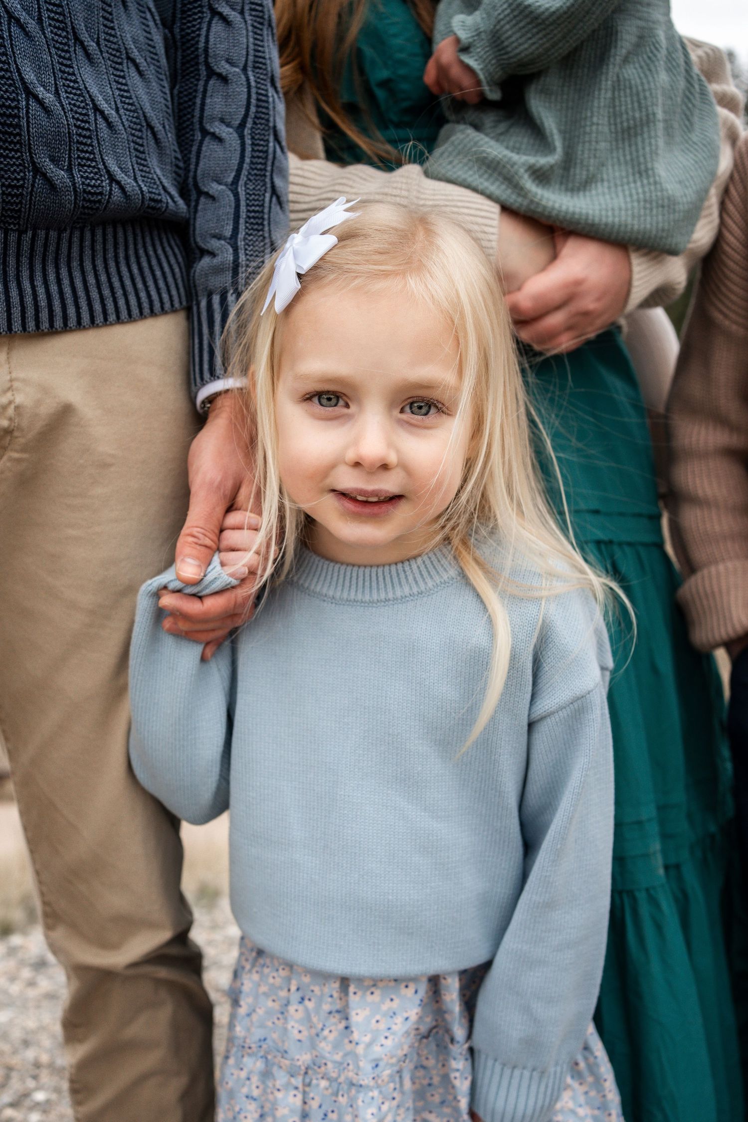 A child wearing a light blue sweater and patterned skirt smiles while standing outdoors.
