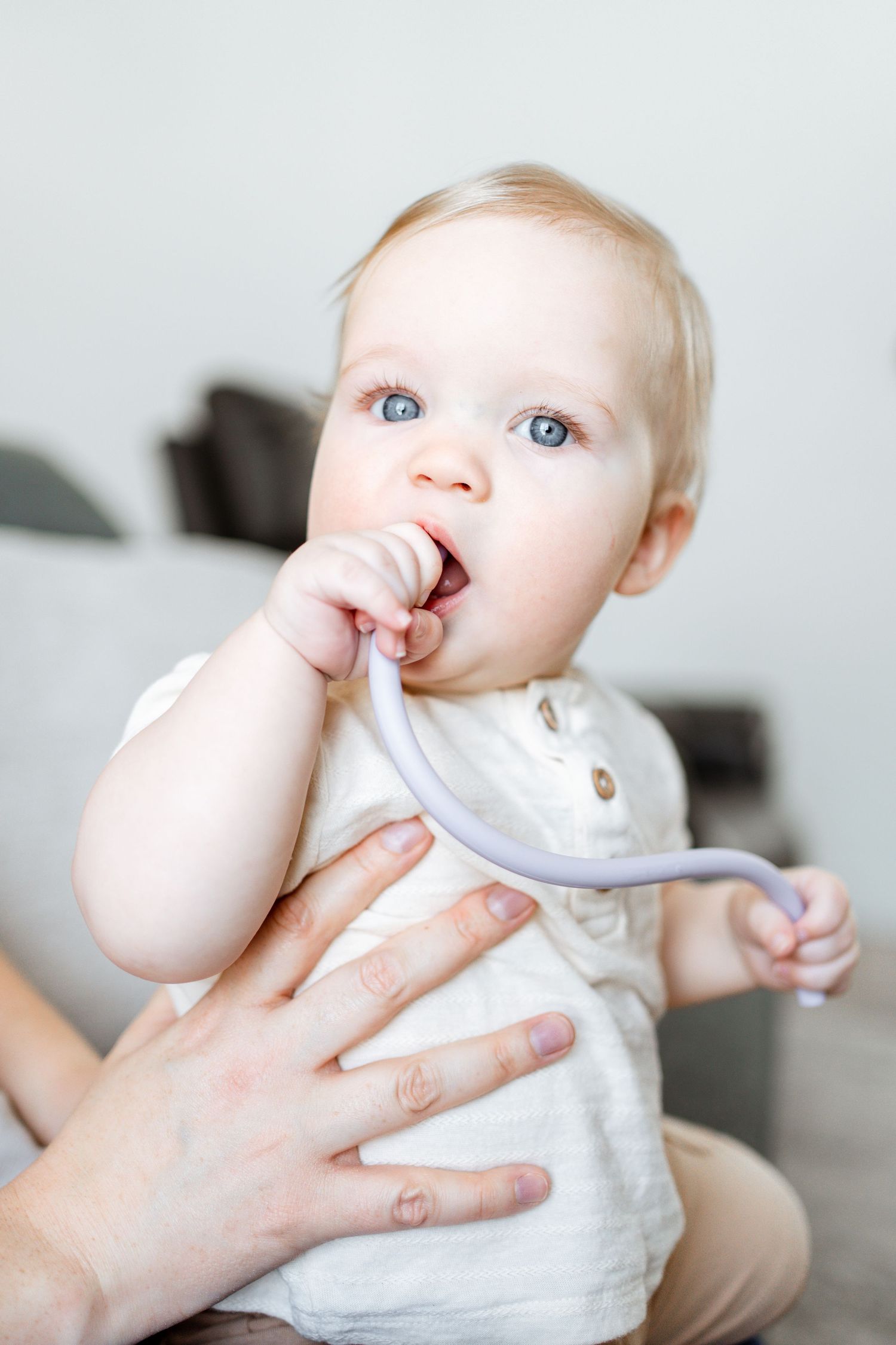 A baby in white clothing touches their nose while being held by adult hands in a bright, minimalist setting.