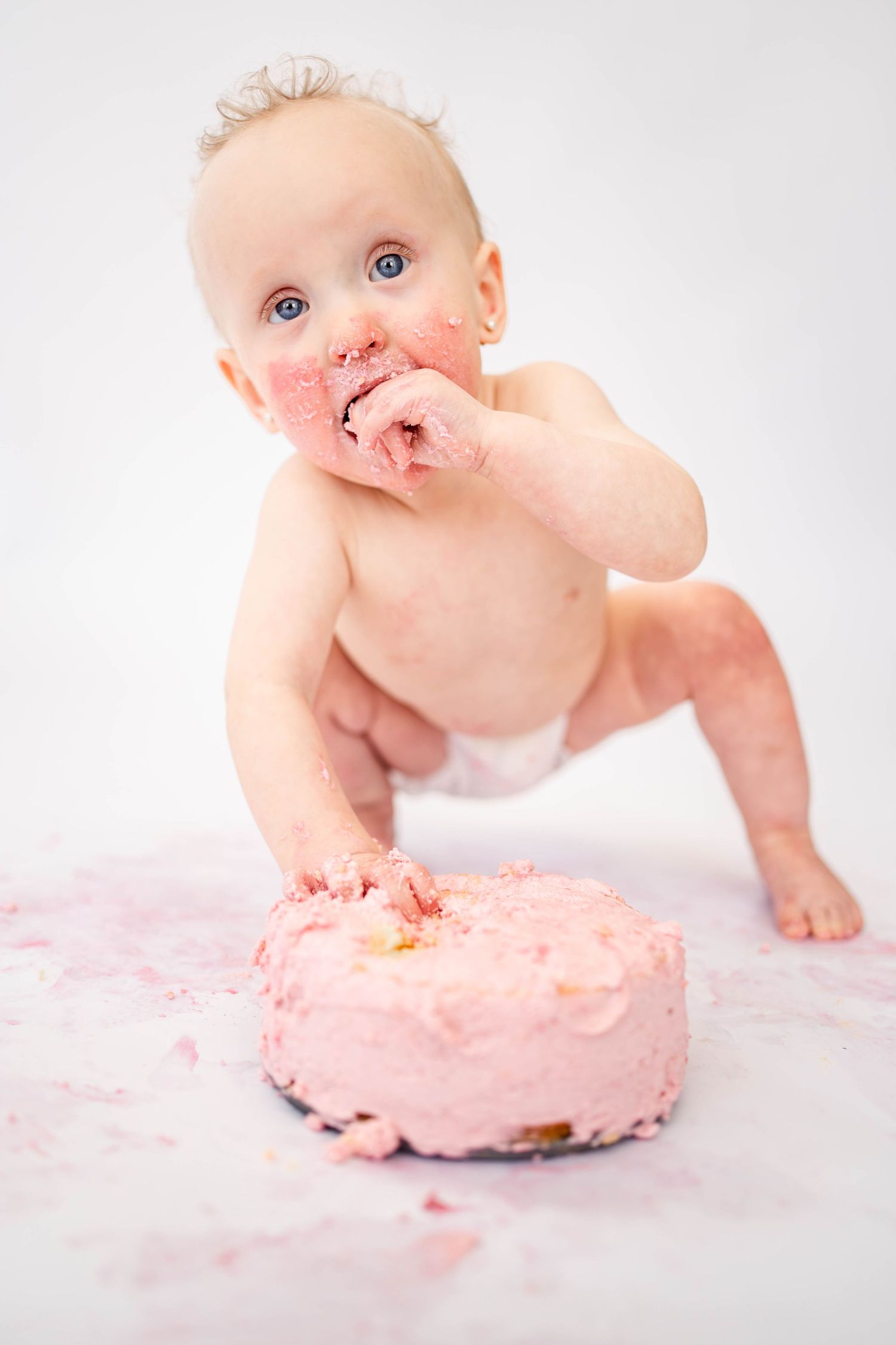 Baby with blue eyes smashing a pink birthday cake, covered in frosting, during a first birthday cake smash photo shoot.