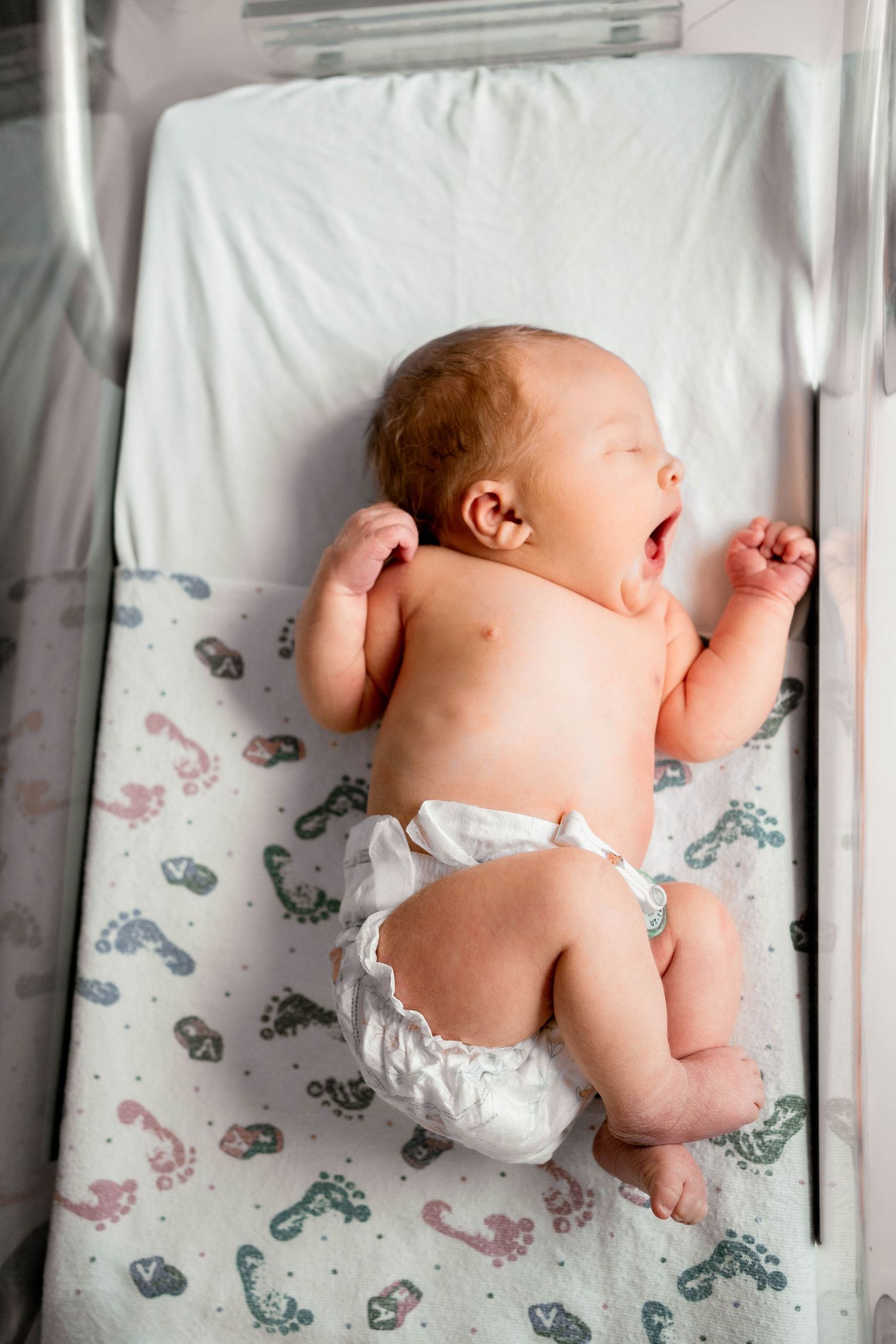 Newborn baby in diaper lying on back in hospital bassinet, yawning with arms raised on footprint patterned blanket.