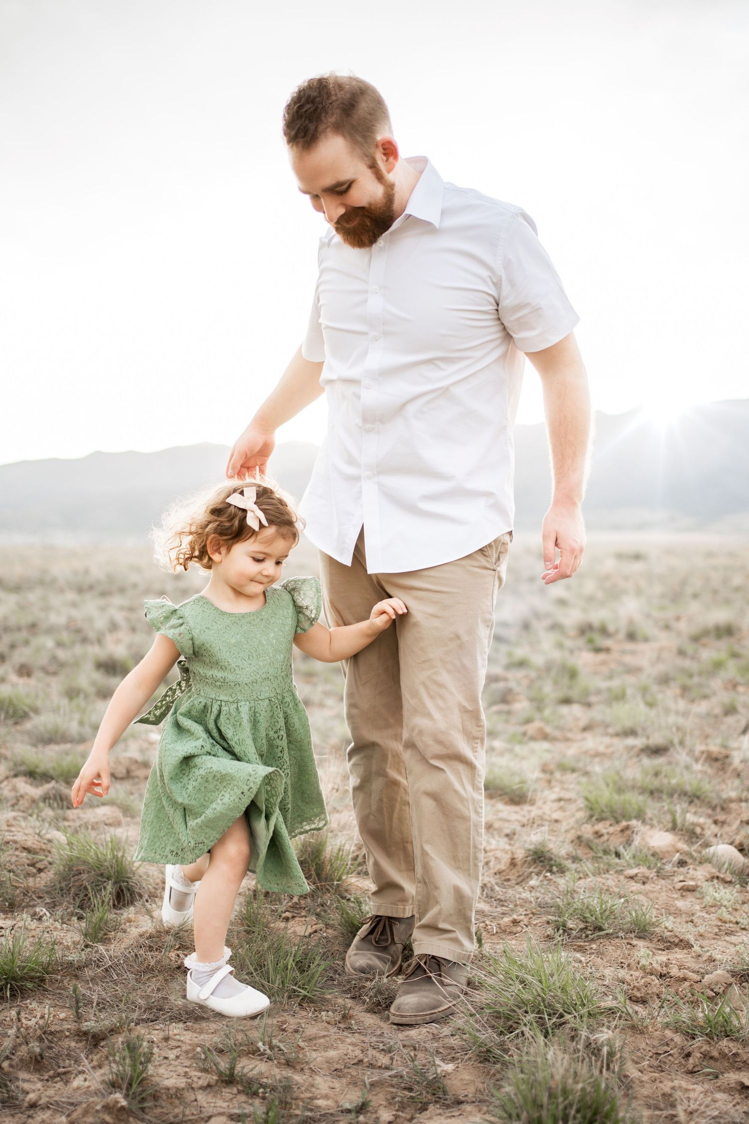 A bearded father in a white shirt holds his young daughter's hand as they walk together in an open field.
