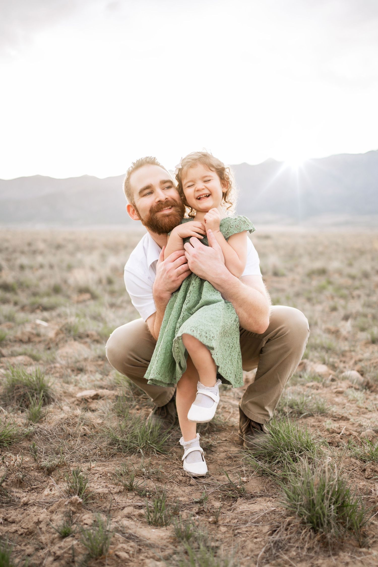 Bearded father crouching in a field, joyfully hugging his laughing little girl in a green dress.