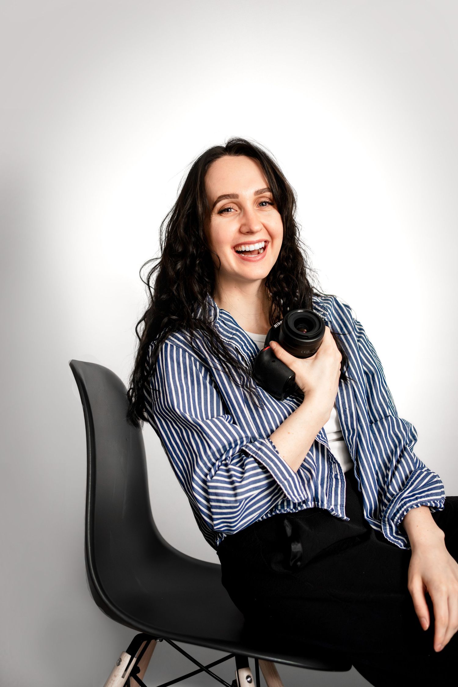 Professional photographer in striped shirt sits on black chair holding camera and laughing against white background.