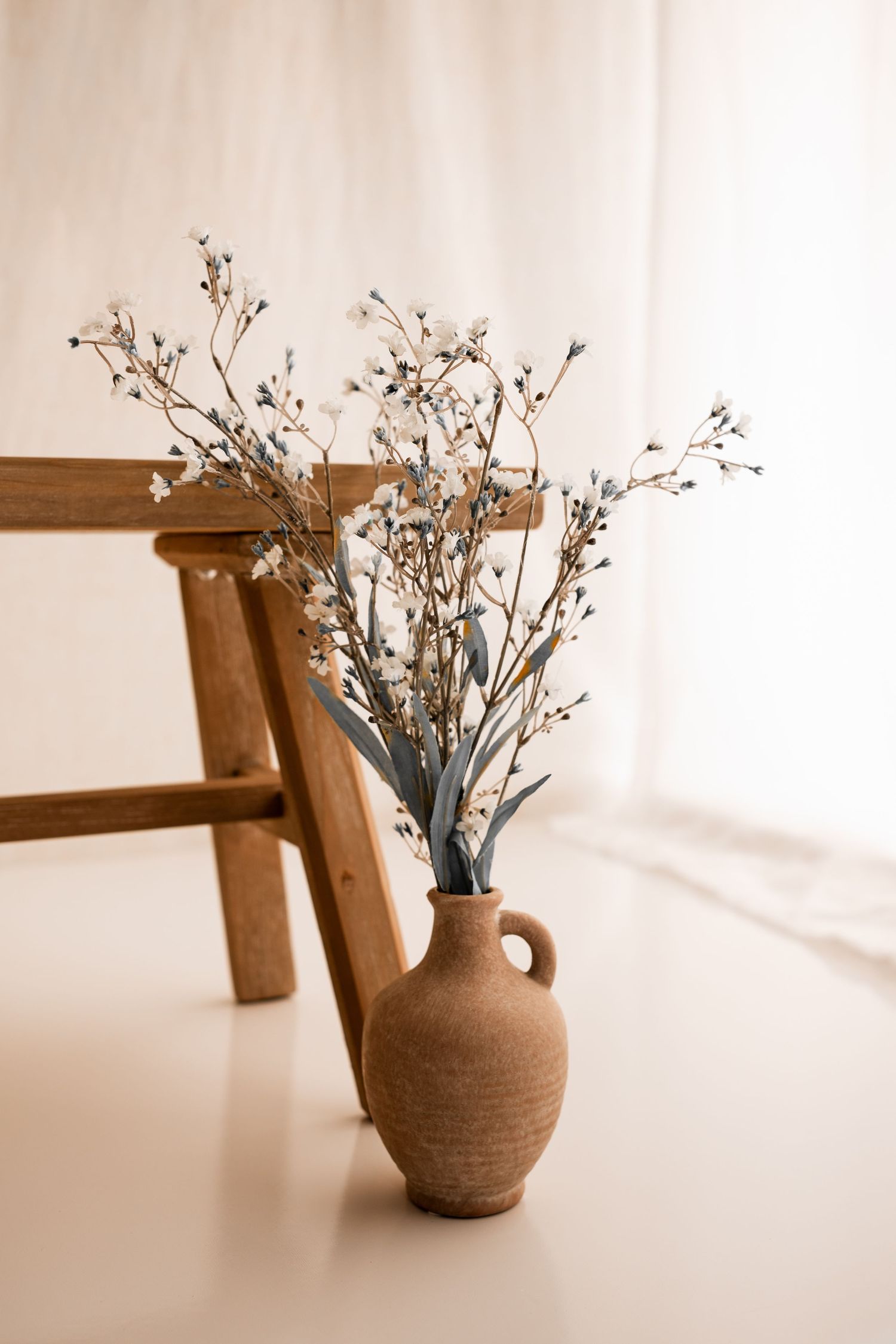 Rustic terracotta vase with dried white baby's breath and eucalyptus stems beside a wooden stool against a soft white curtain.
