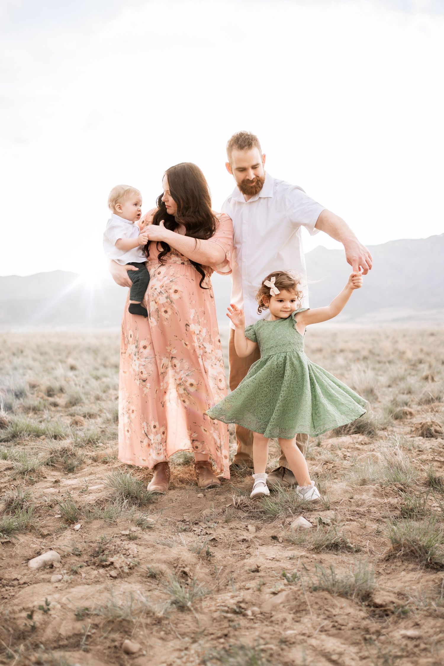 Family of four with mother in floral dress and father dancing with toddler in green dress outdoors.