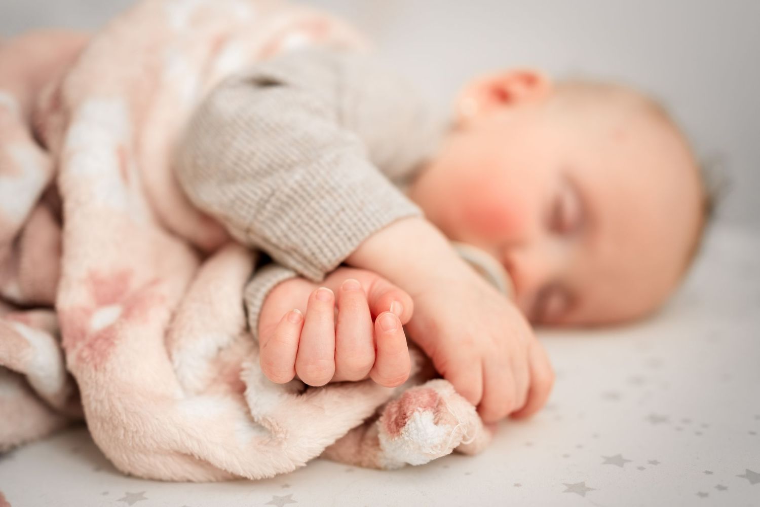 A tiny hand grasps a soft beige knit fabric while sleeping peacefully on a light textured surface.