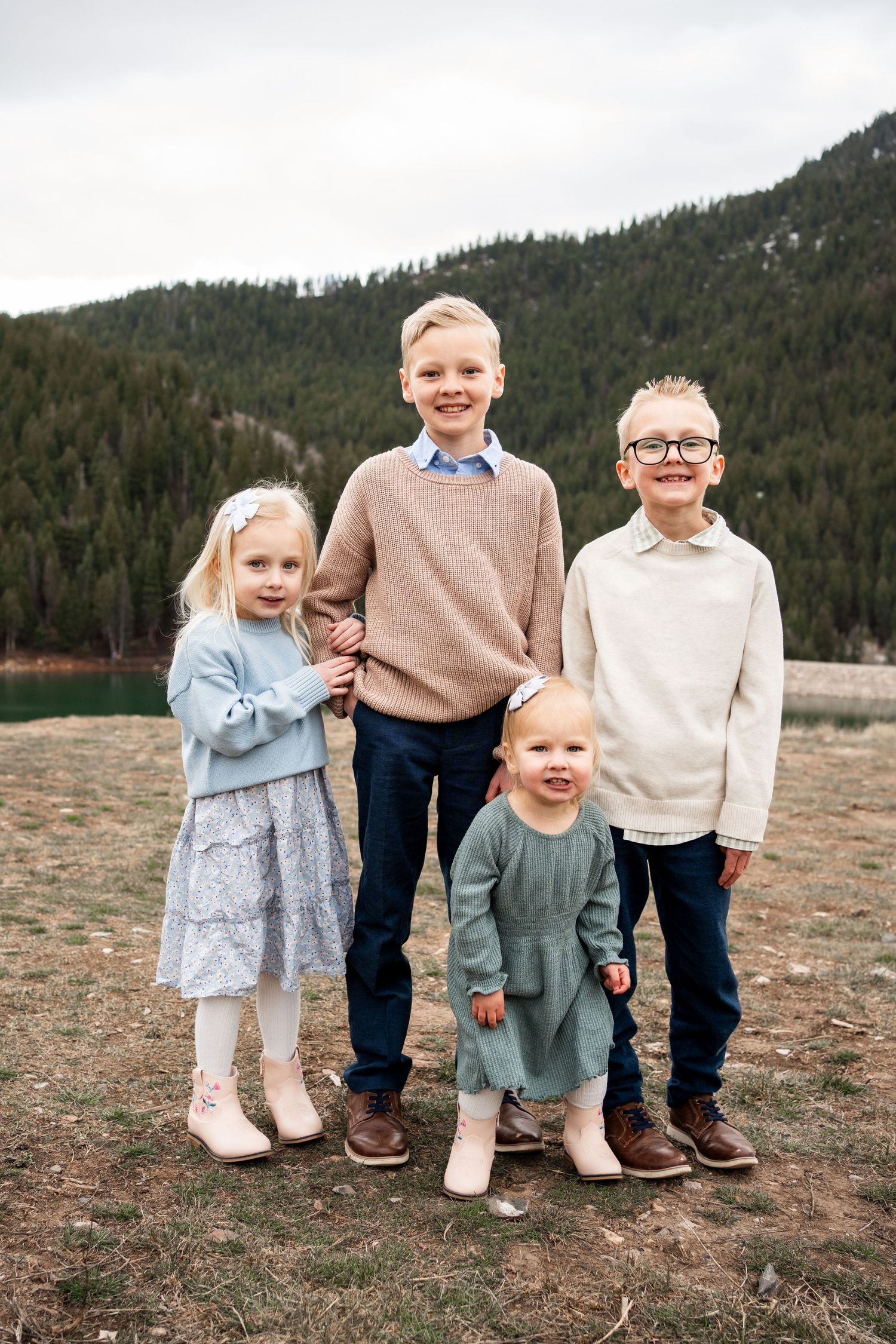 Children's feet and legs shown in casual clothing standing together outdoors with mountains and forest in background.