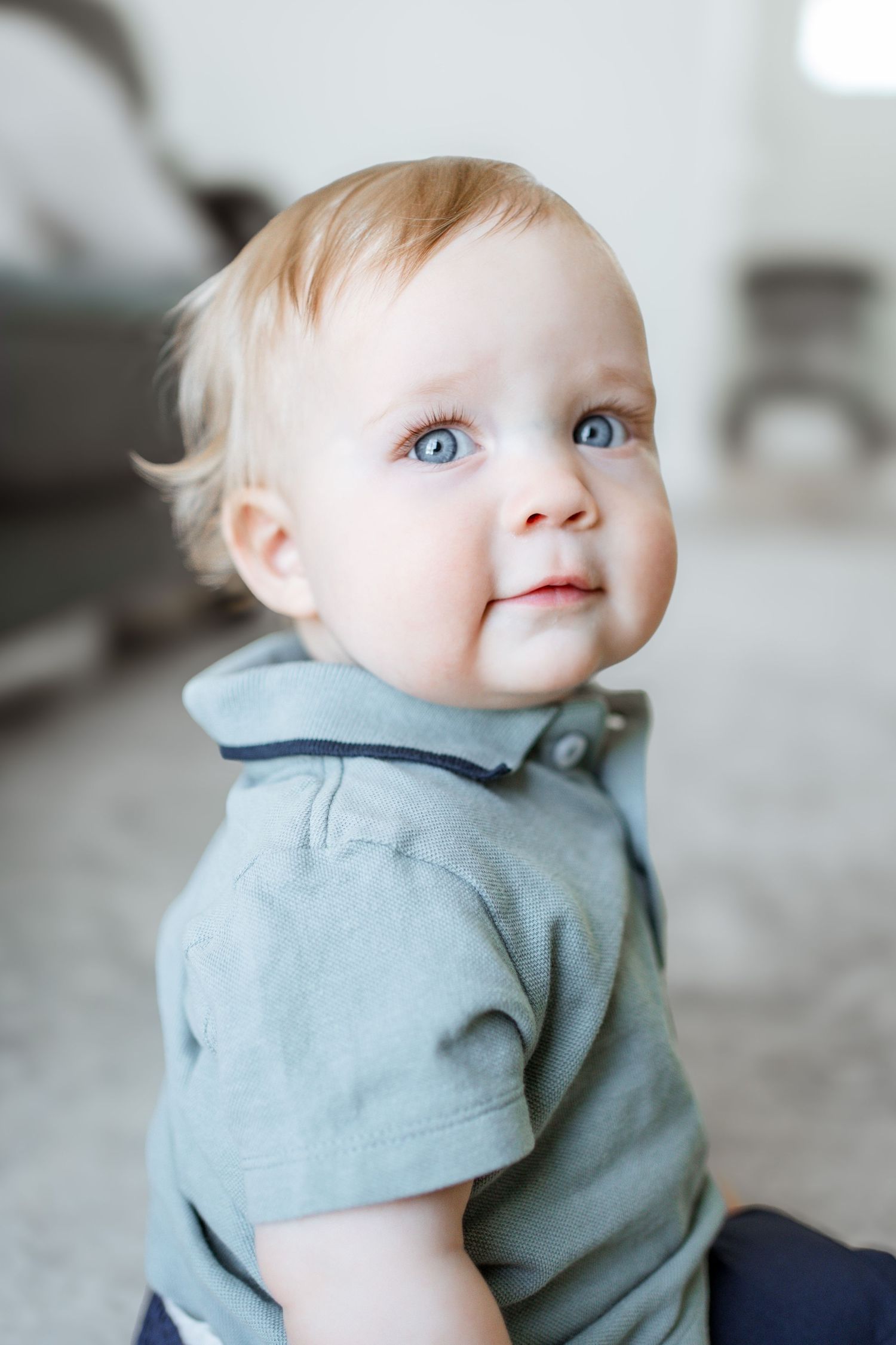 Baby in soft grey top looks up with bright blue eyes against neutral background.