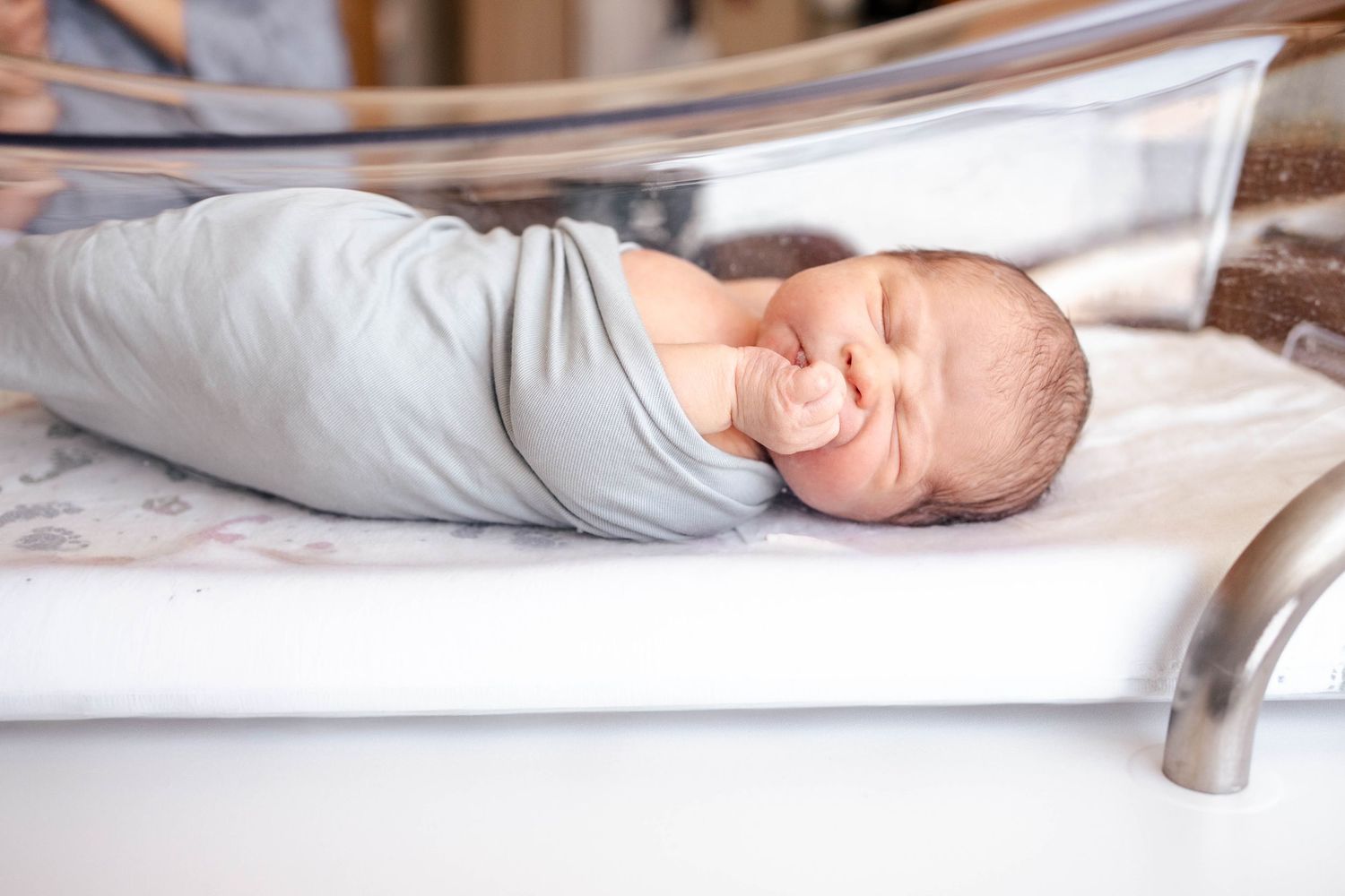 Newborn baby swaddled in gray blanket sleeping peacefully in hospital bassinet.