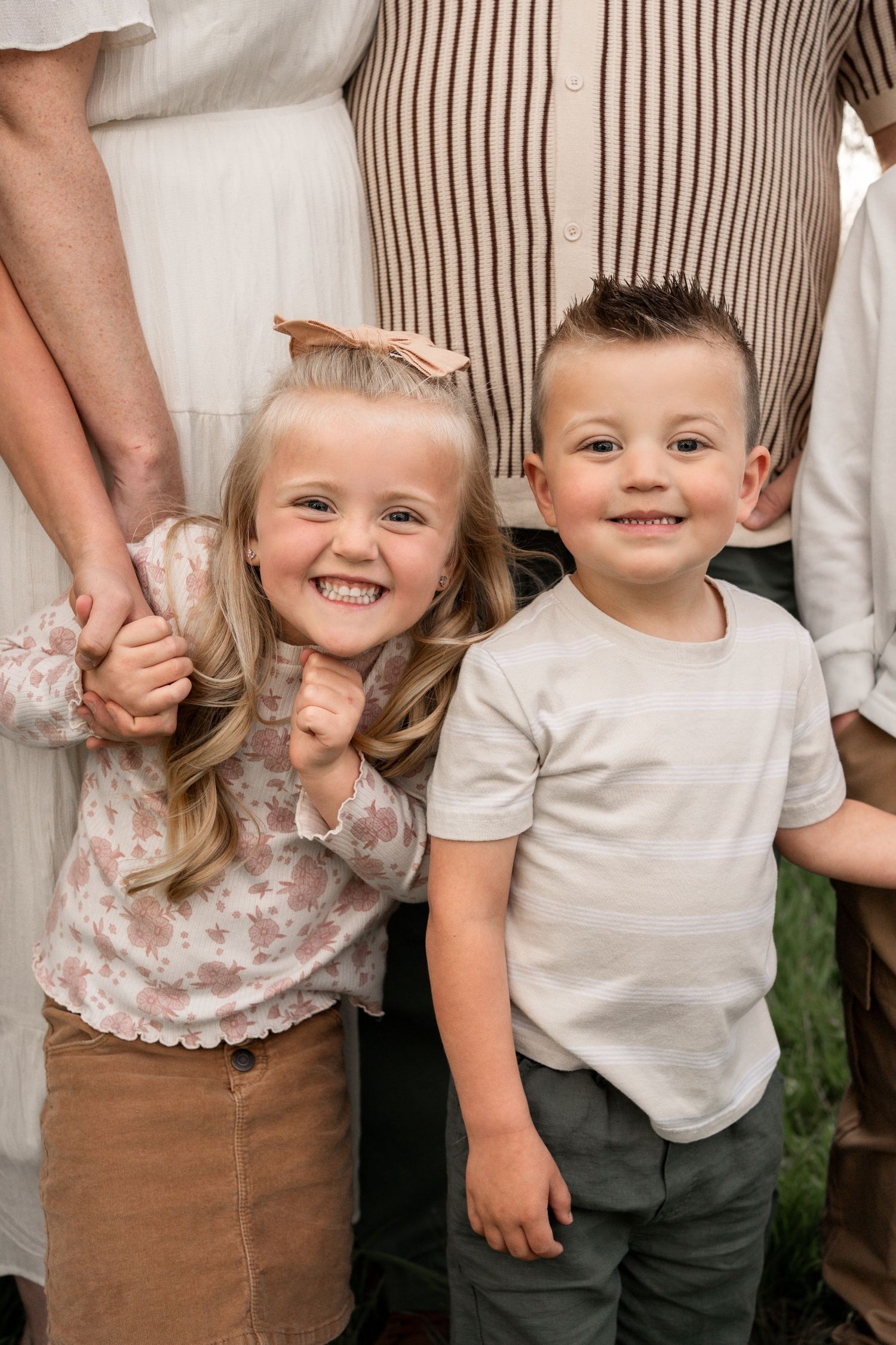 Two smiling young children, a girl with a bow and a boy, stand together in a warm family portrait photo.