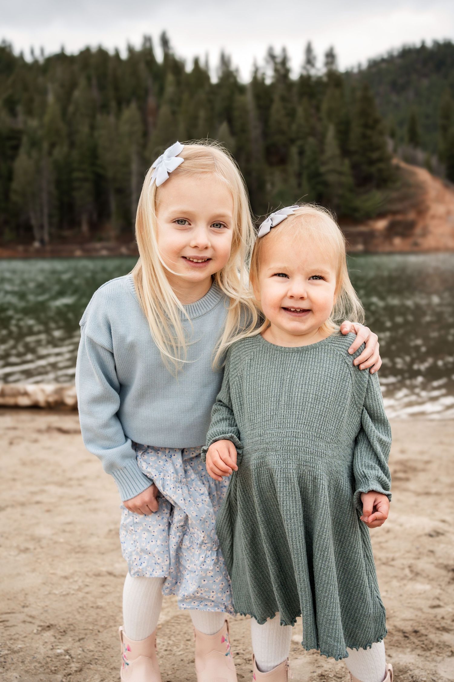 Two young children in knit sweaters and hair bows smile together by a lake.