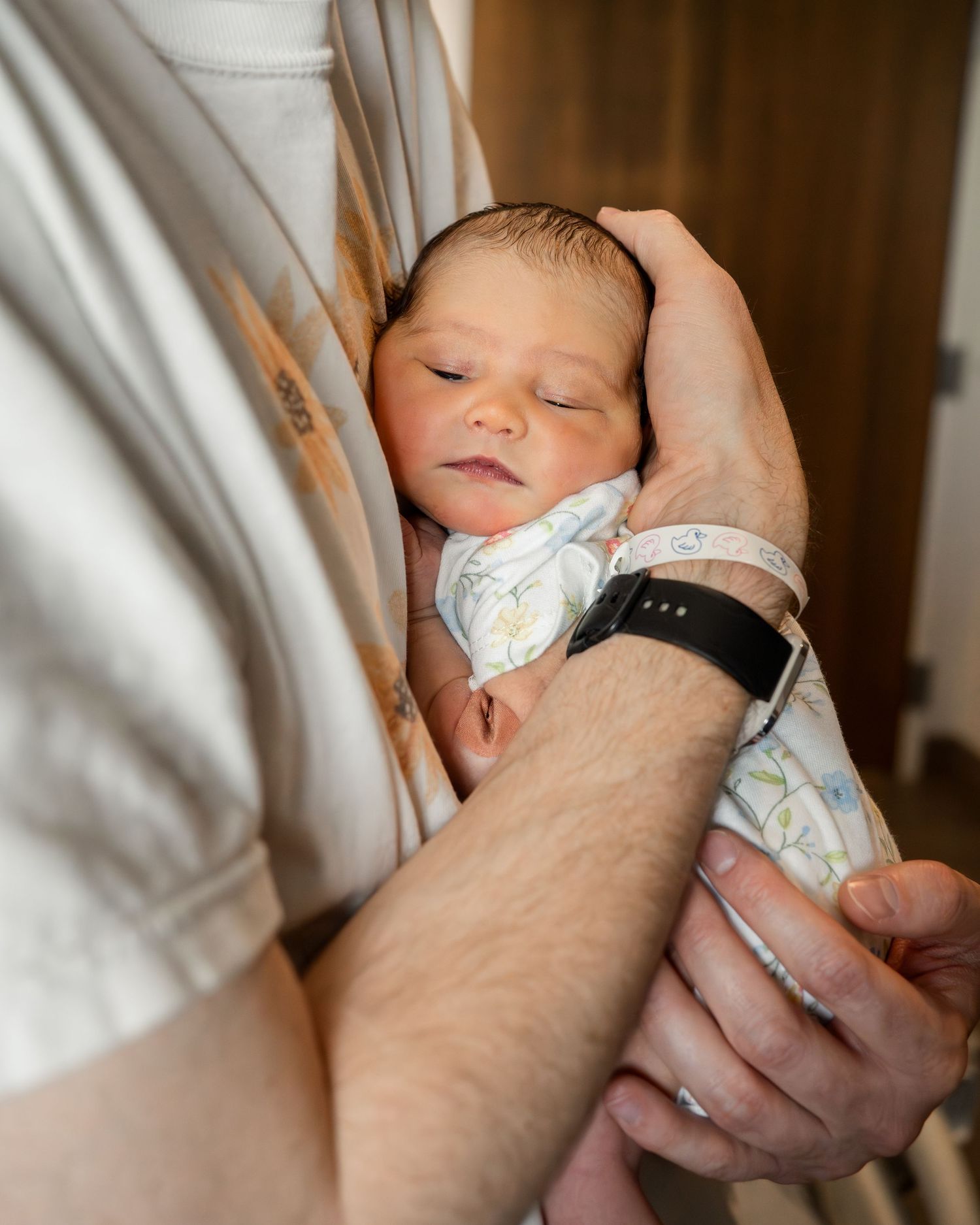 Father cradling a sleeping newborn baby in his arms while wearing a smartwatch in a hospital room.