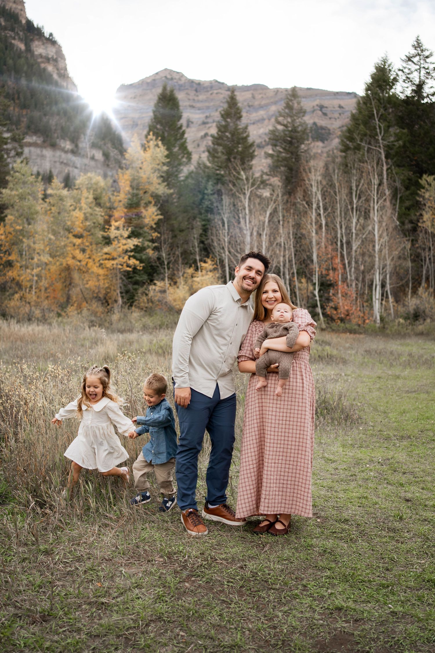 Family of five in coordinated neutral outfits poses on a grassy mountain meadow with autumn trees and sunlight behind them.