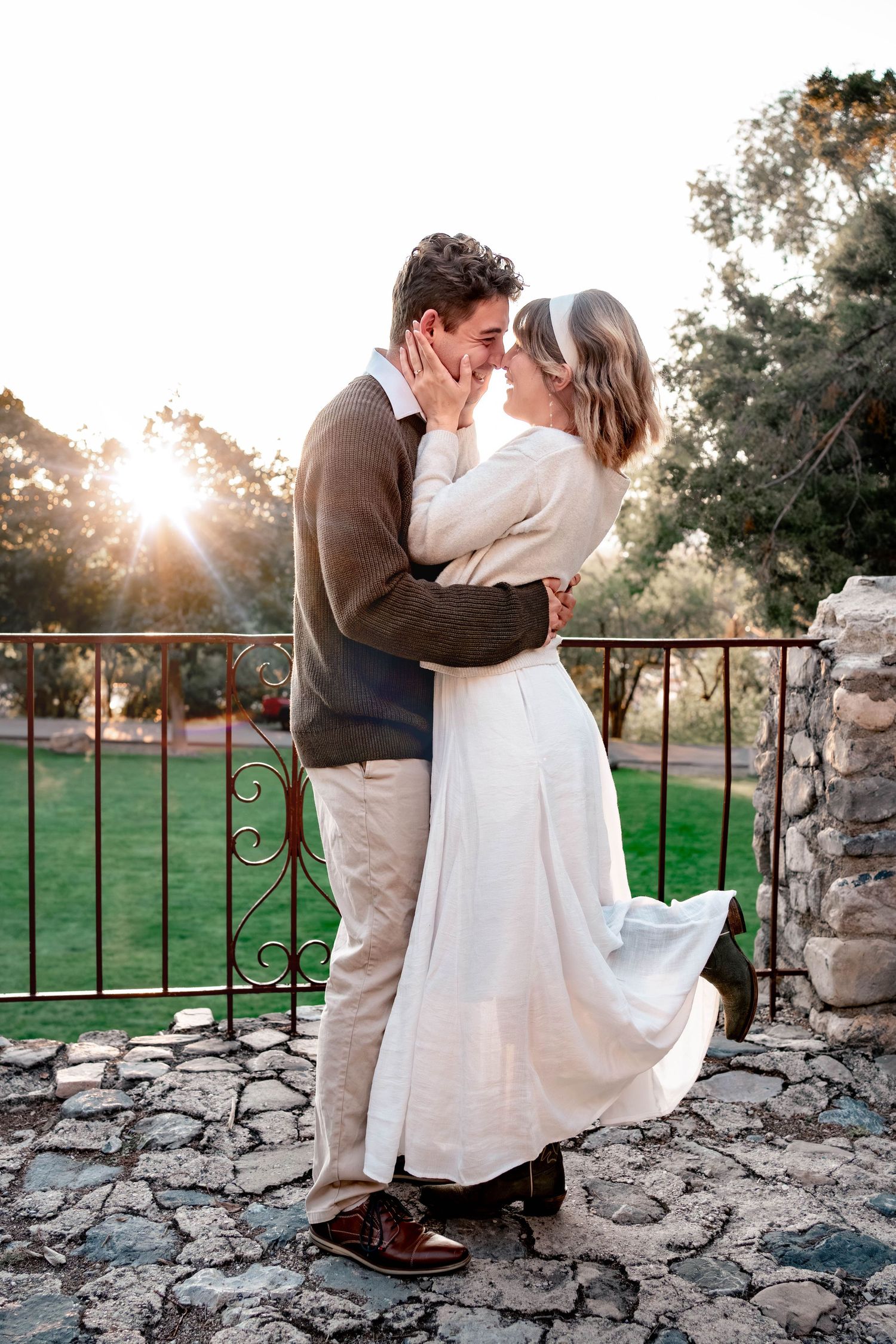 Couple embracing and touching foreheads on a stone patio at sunset, with a green lawn, iron railing and trees glowing in warm golden light.