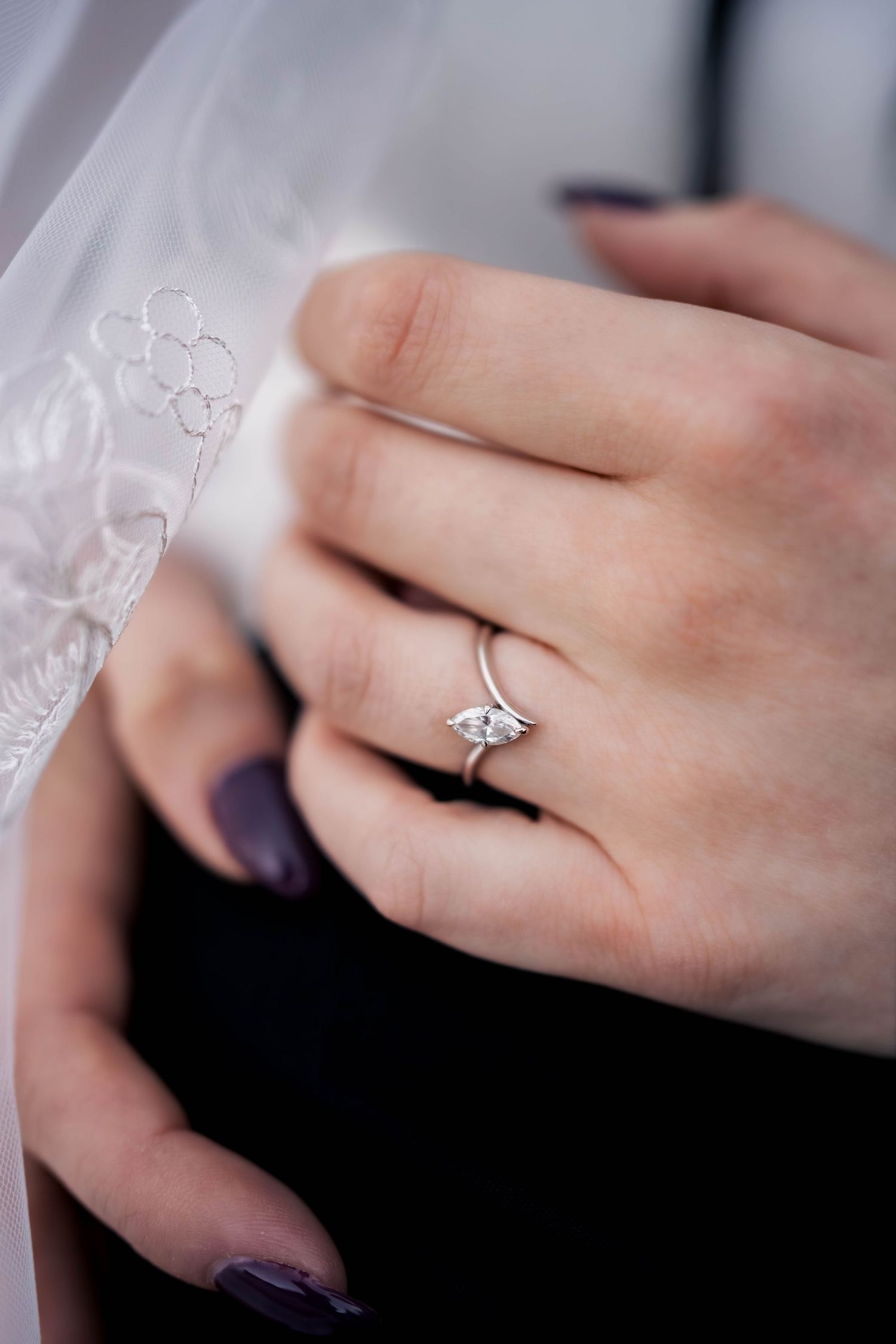 Close-up of a bride's hand with dark purple nails wearing a marquise diamond engagement ring beside a lace-embroidered veil.