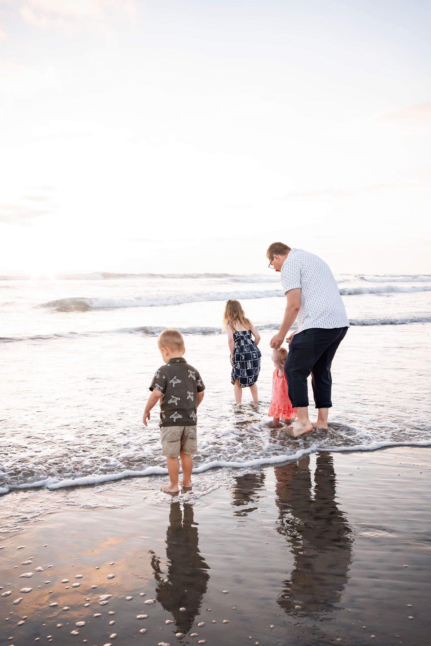 Family of four wading in ocean waves at sunset on a sandy beach, with children and adult enjoying the shoreline together.