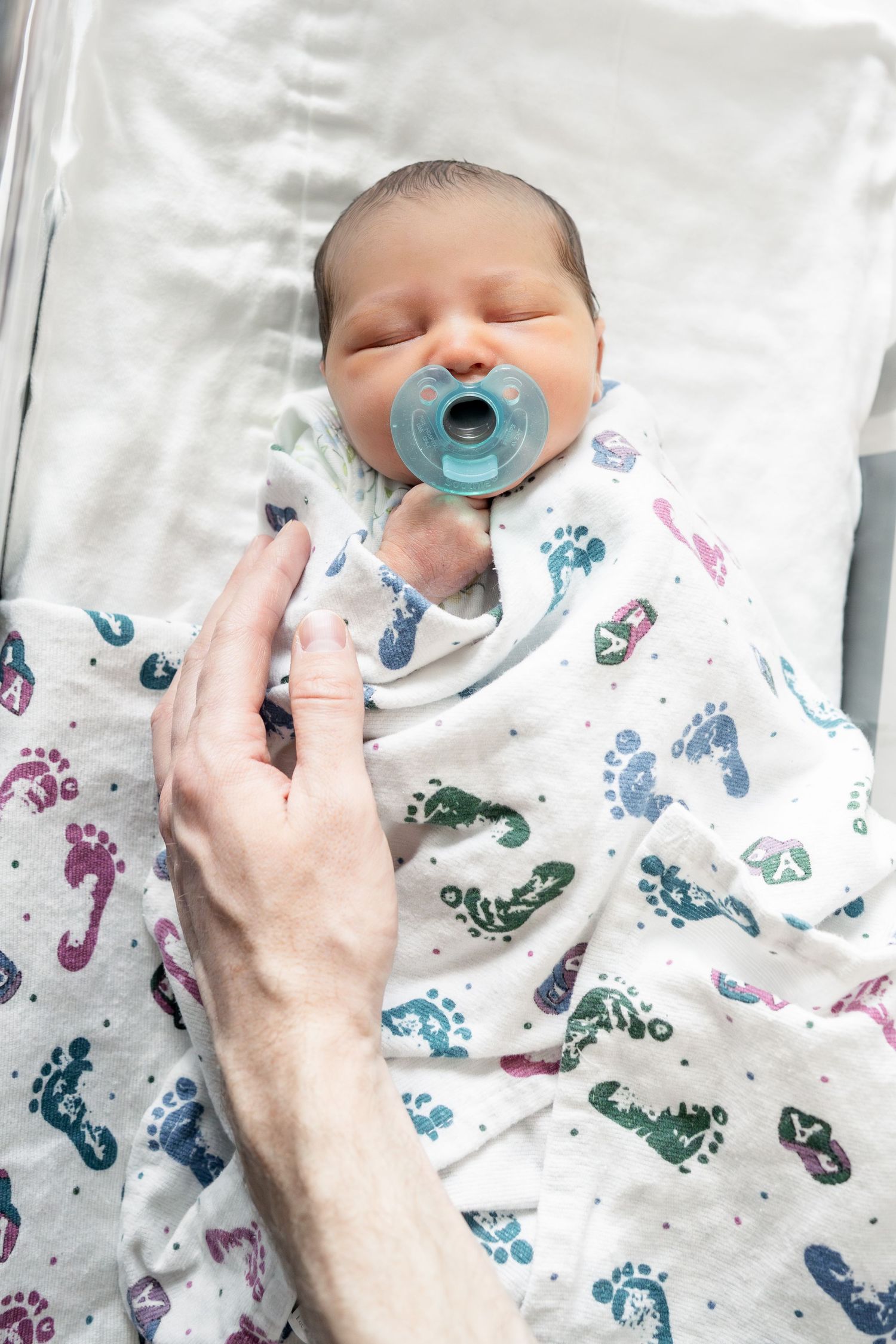 Newborn baby swaddled in footprint blanket with blue pacifier, resting in hospital bassinet with adult hand nearby.