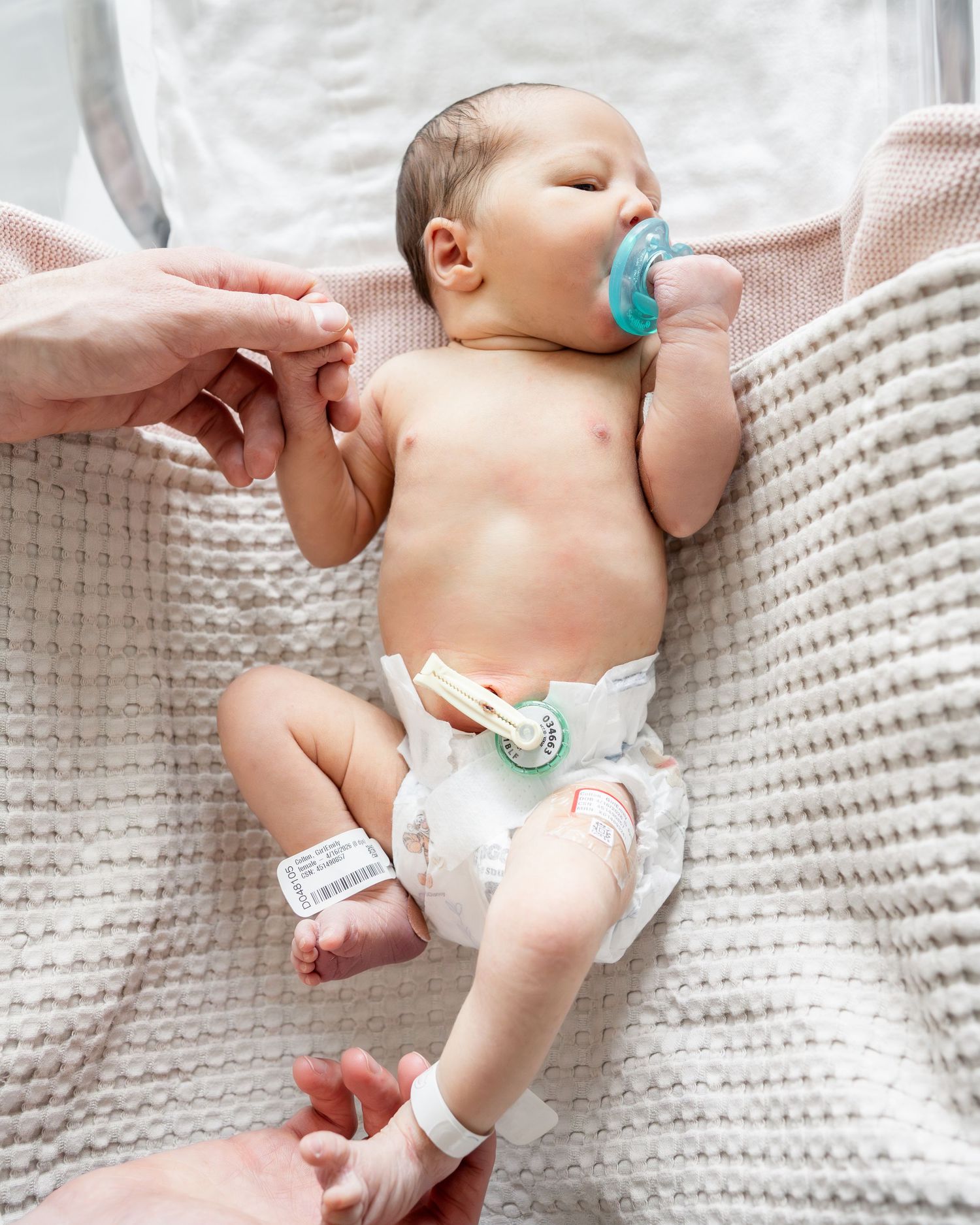 Newborn baby lying on white knit blanket with pacifier, wearing diaper and hospital bracelet, held by adult hands.