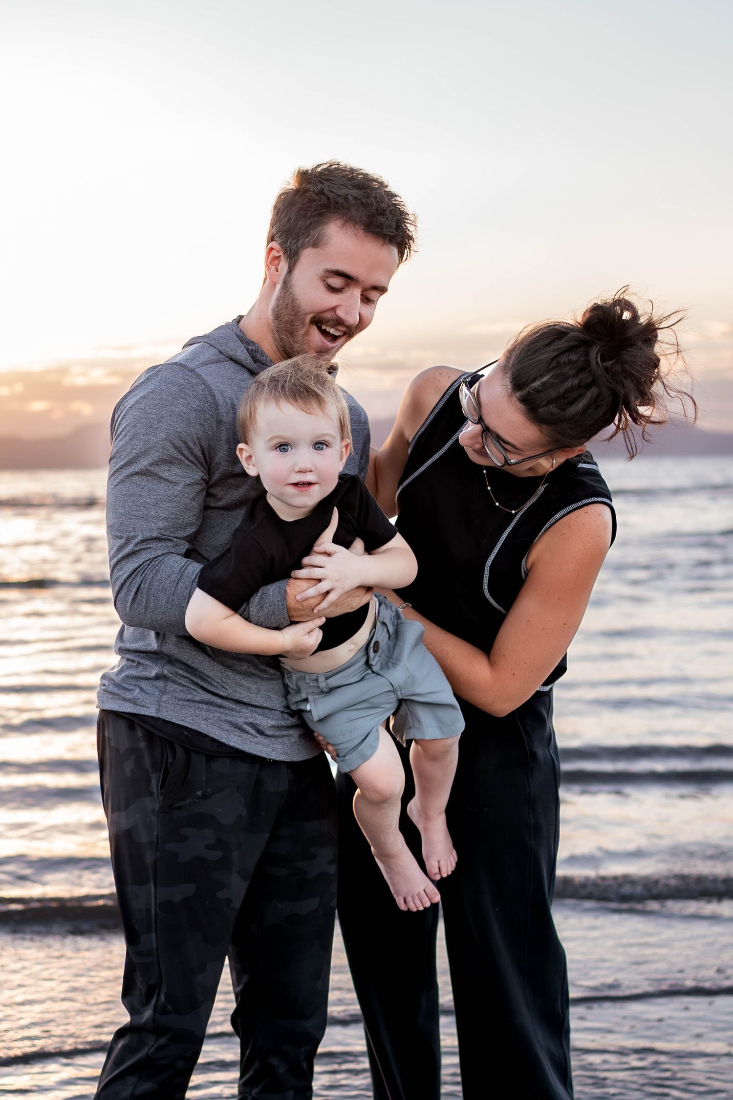 A smiling couple holds a laughing toddler between them on a sandy beach at sunset, with calm ocean water behind them.