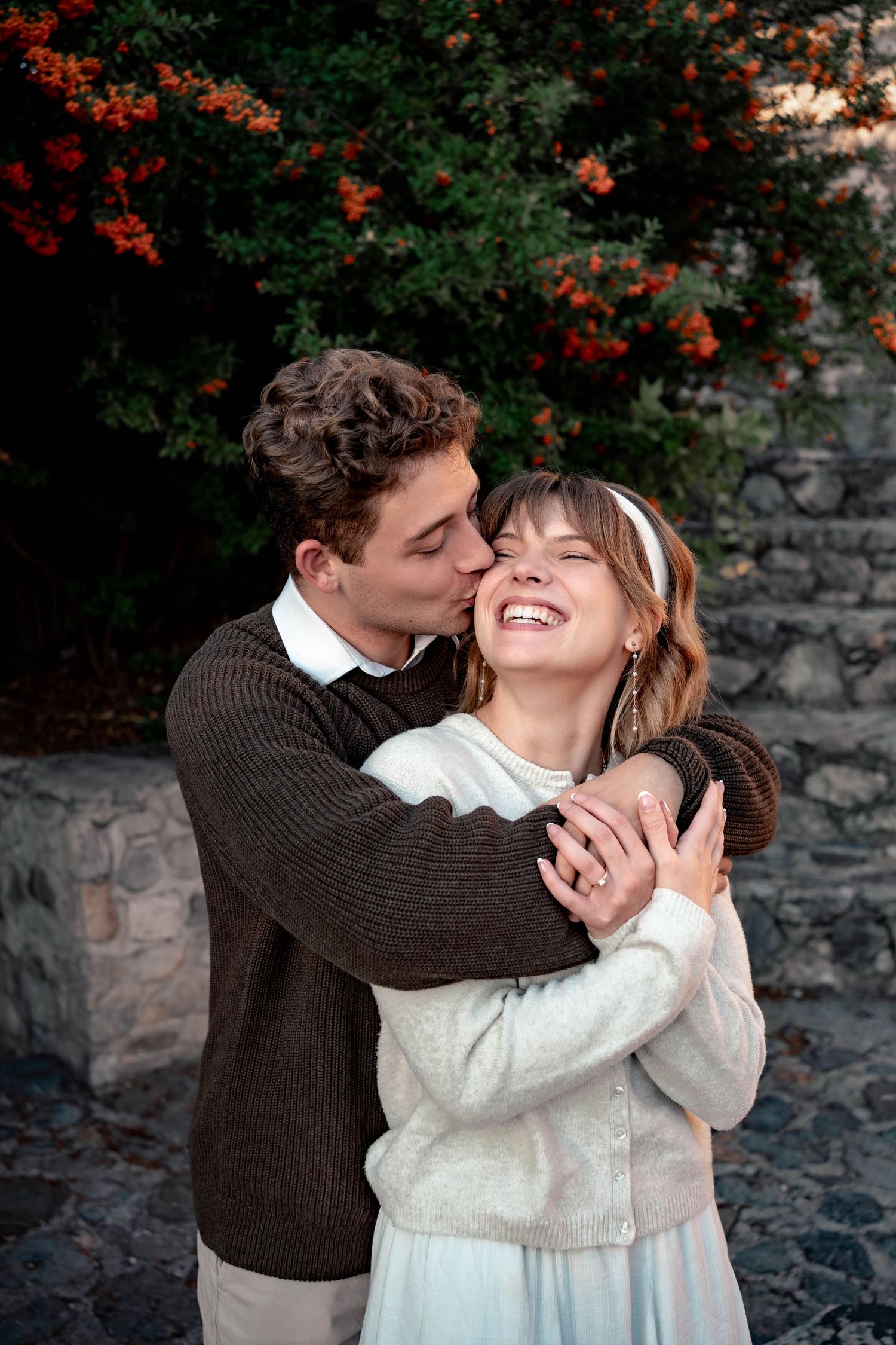 Smiling young woman in a white cardigan and headband is embraced and kissed on the cheek by a man in a brown sweater near a stone wall.