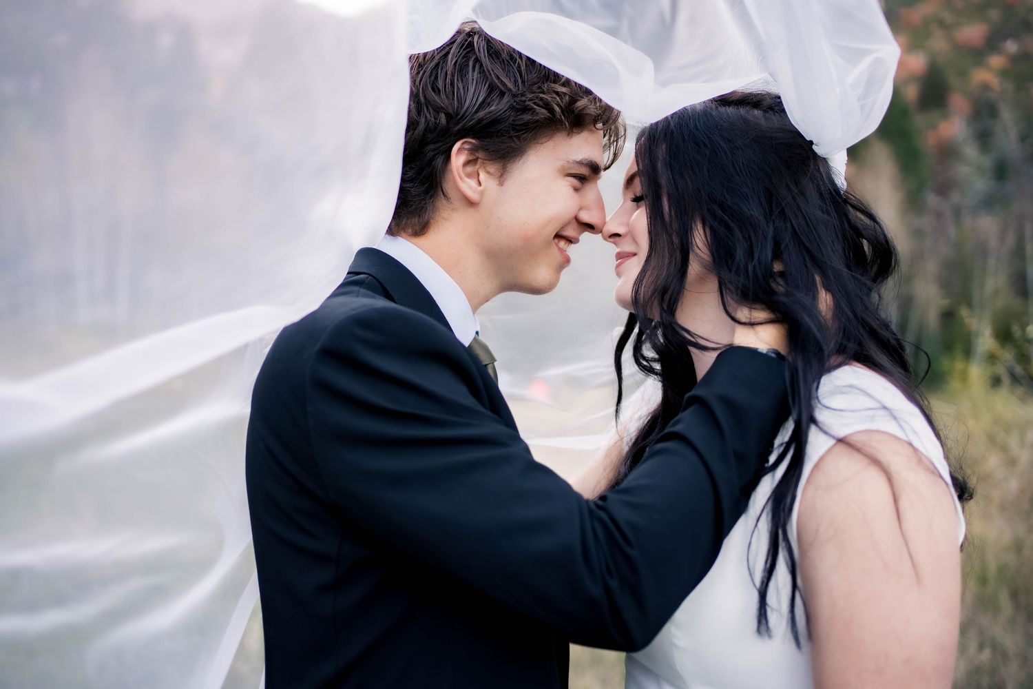 Smiling young couple in formal wedding attire share a tender moment, foreheads touching, beneath a flowing sheer white veil outdoors.