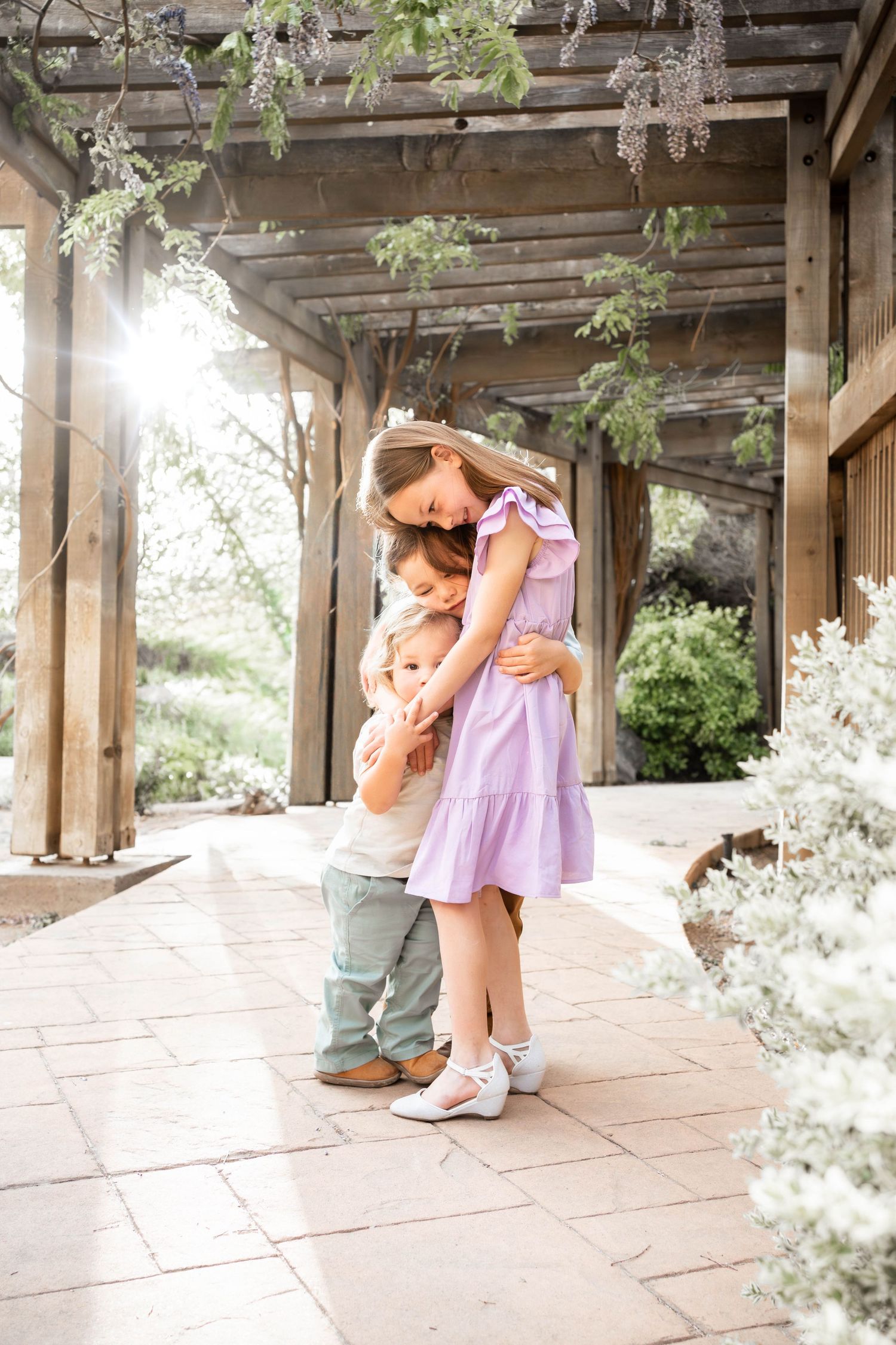 Two children share a sweet hug under a wooden pergola surrounded by blooming flowers in soft natural light.