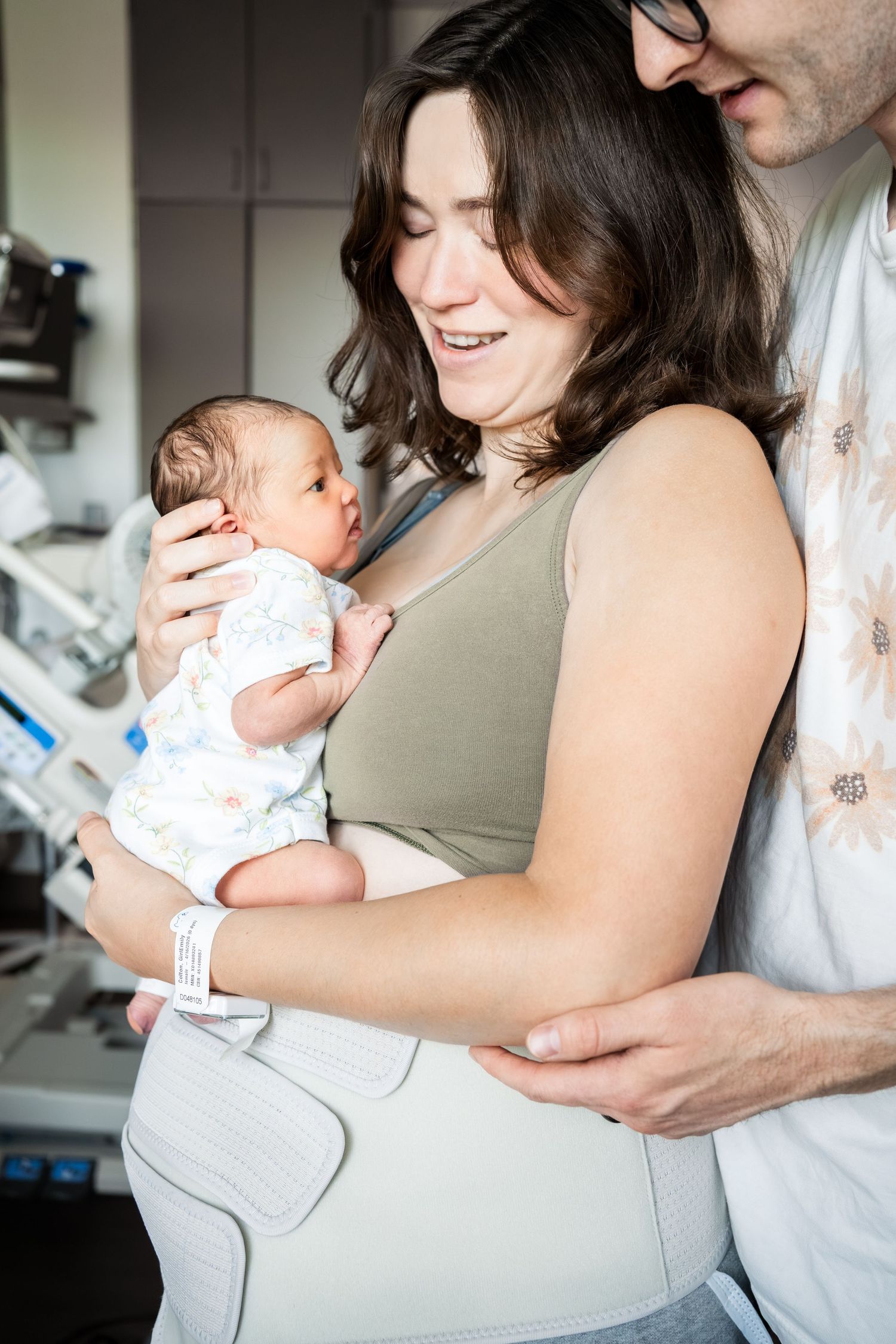Happy new parents smiling and holding their swaddled newborn baby in hospital room.