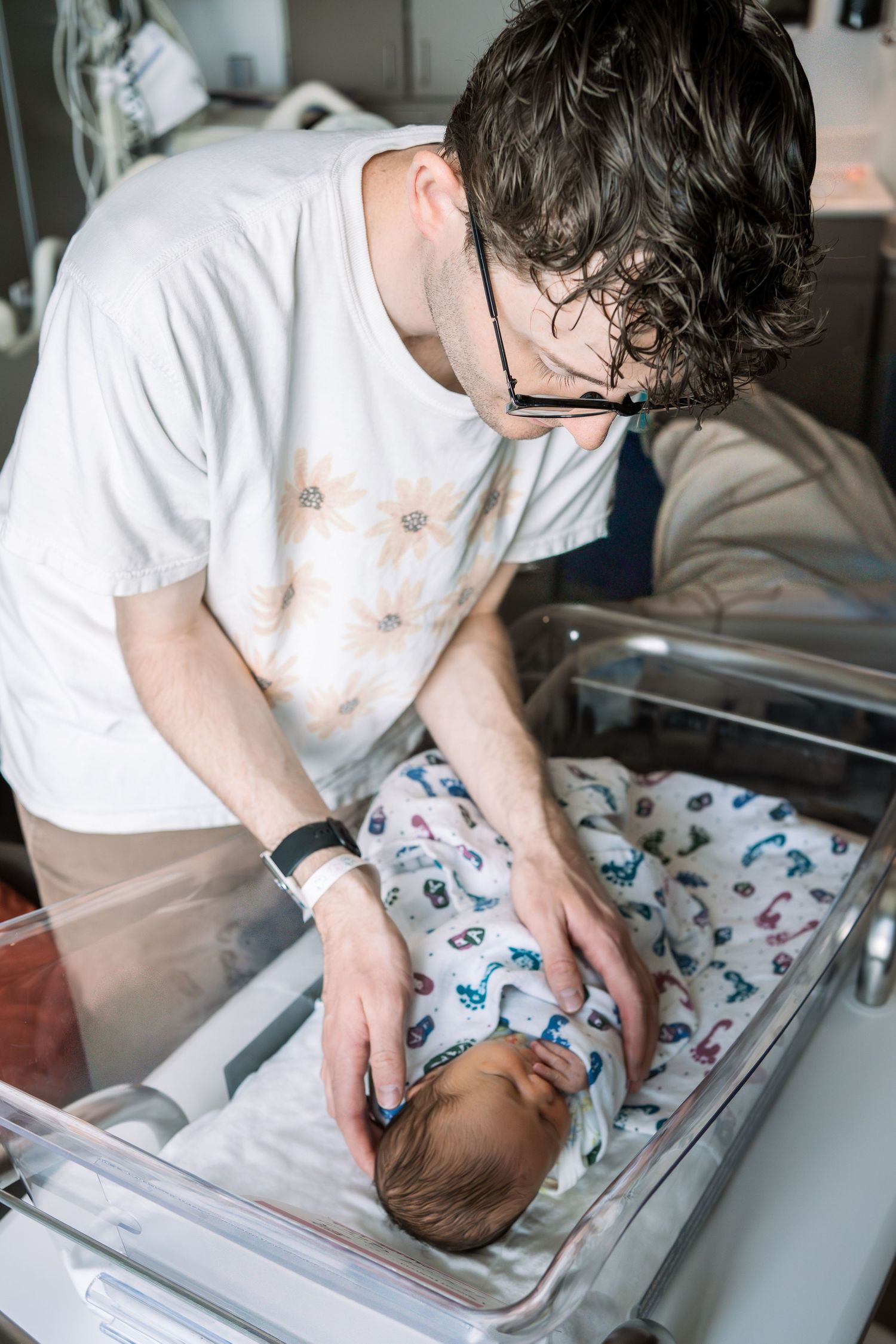 New father with glasses tenderly caring for his newborn baby in a hospital bassinet after birth.