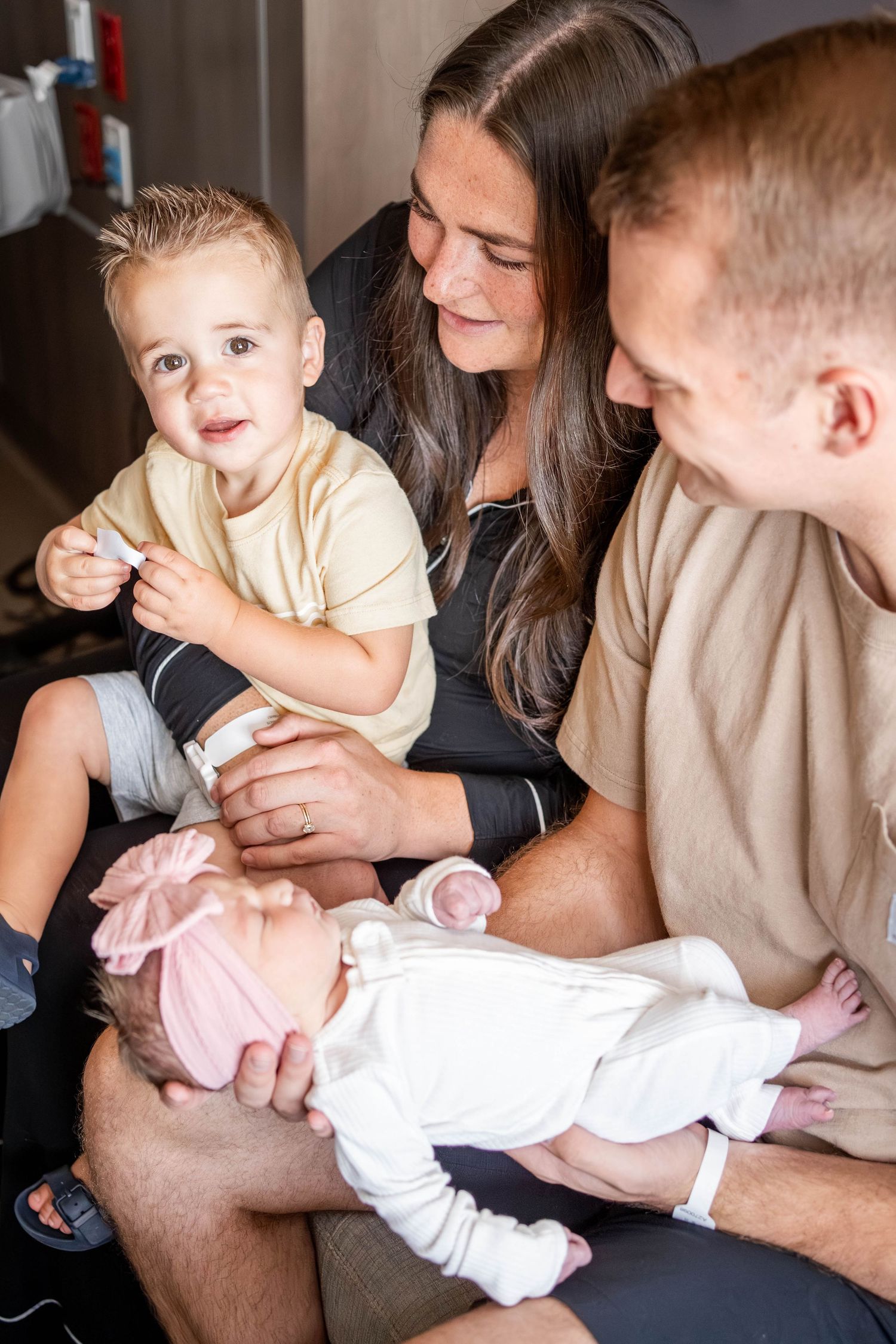 A young family with a toddler boy and newborn baby girl in a pink bow bonnet sitting together indoors.