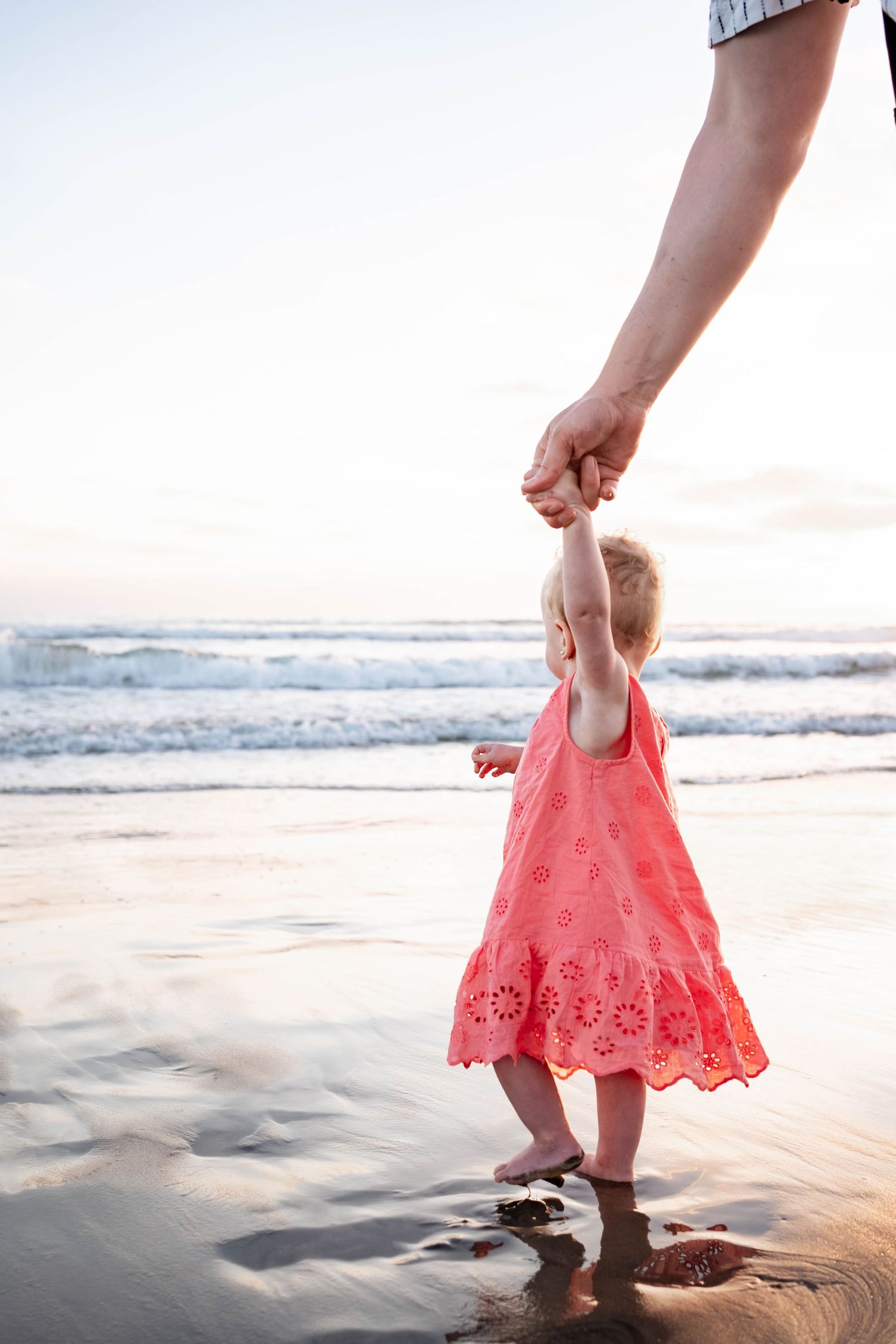 Toddler girl in pink dress holds adult hand while walking barefoot on a sandy beach near ocean waves.