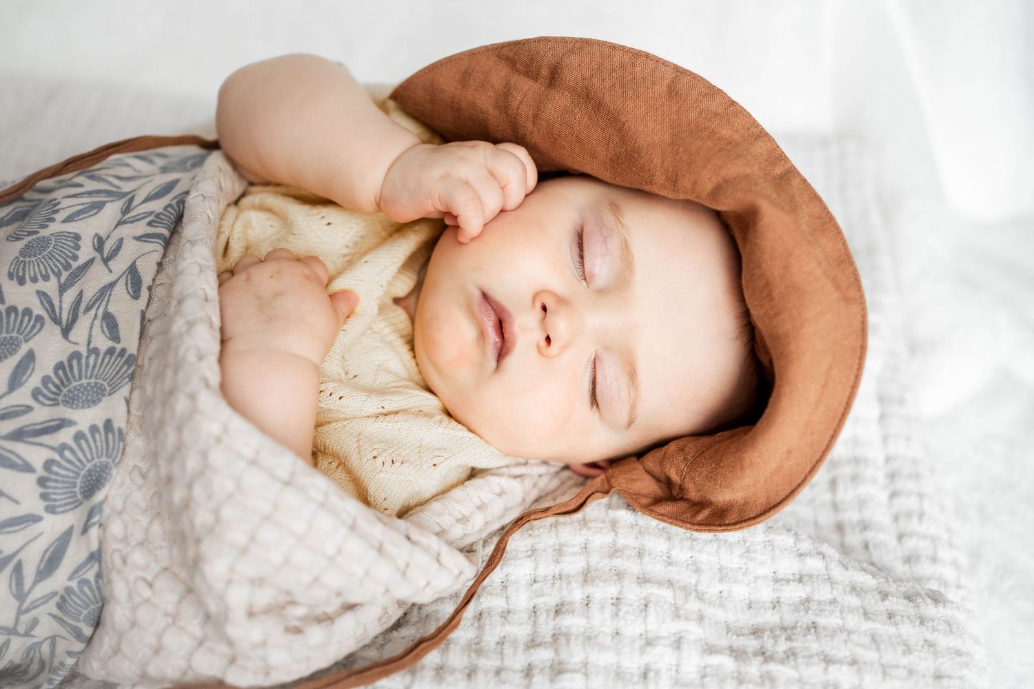 A peaceful newborn baby sleeping in a brown bowl, wrapped in soft white floral blankets, wearing a cozy knit outfit.