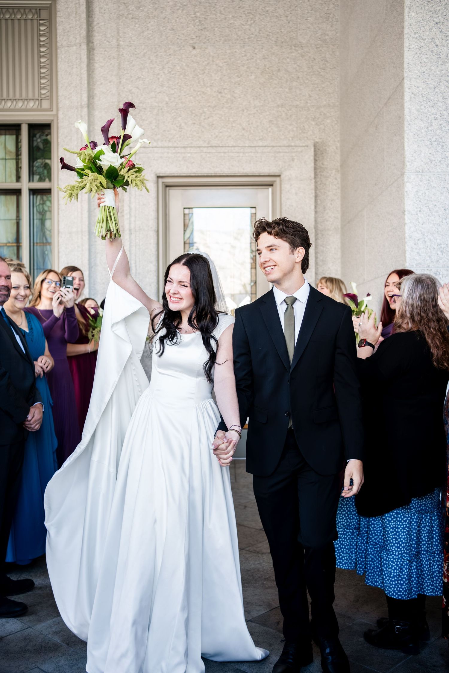 Smiling bride in a white gown raises her bouquet of calla lilies while holding hands with the groom outside as wedding guests cheer.