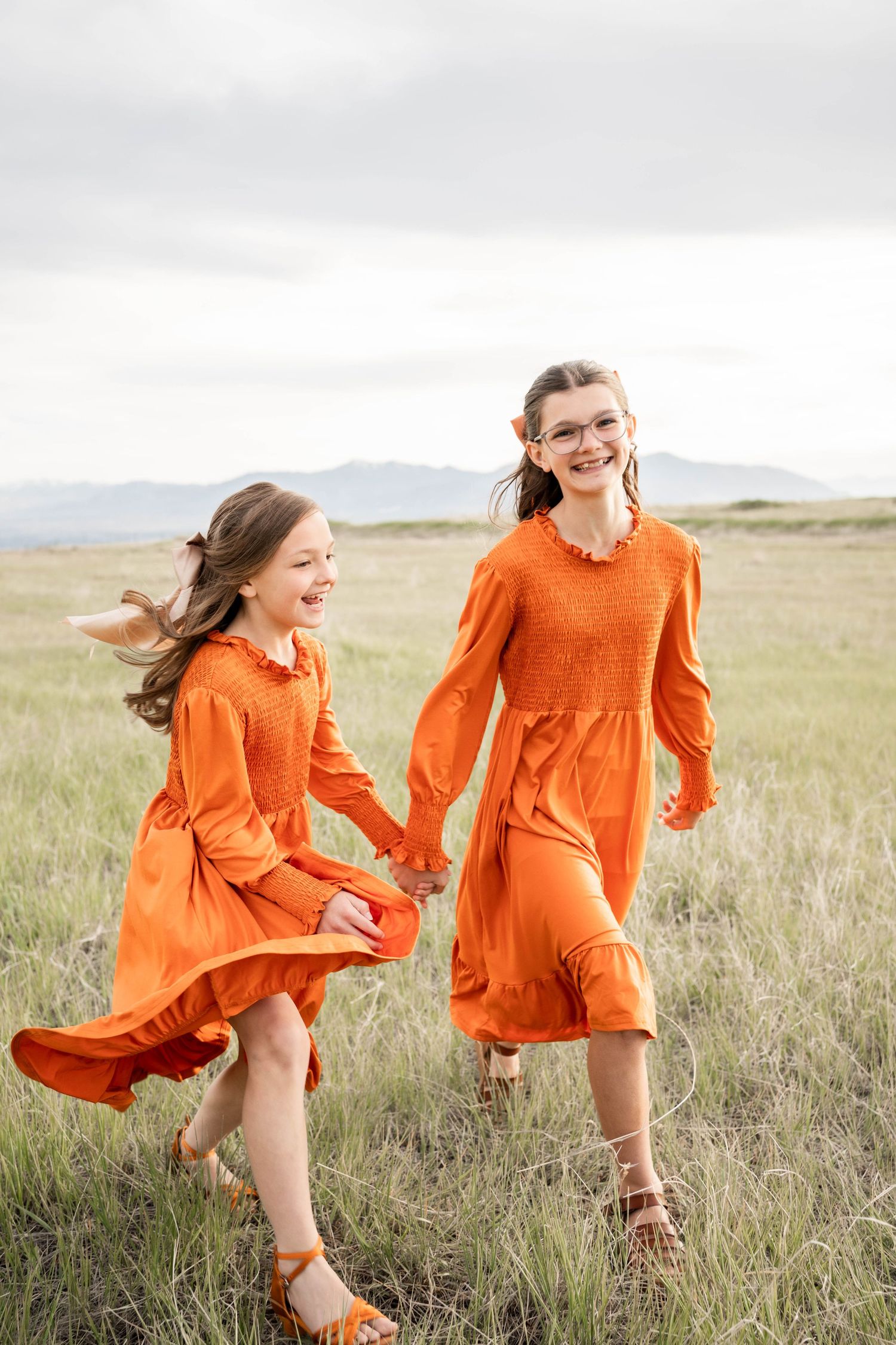 Two girls in matching orange dresses holding hands and laughing while running through a grassy field.