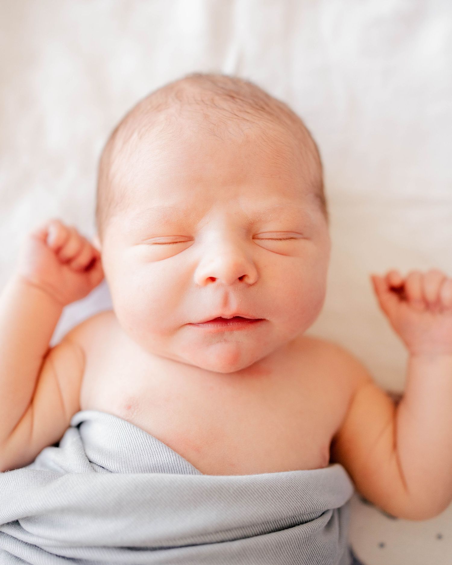 Peaceful sleeping newborn baby wrapped in a soft gray swaddle with tiny fists raised on a white background.