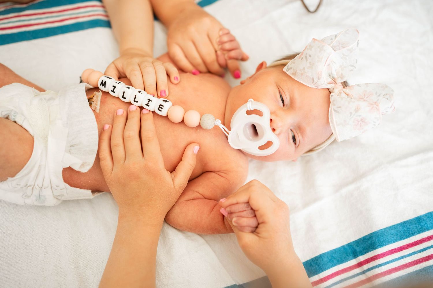 Baby girl with pacifier clip spelling a name lying on white blanket while being dressed by parents.