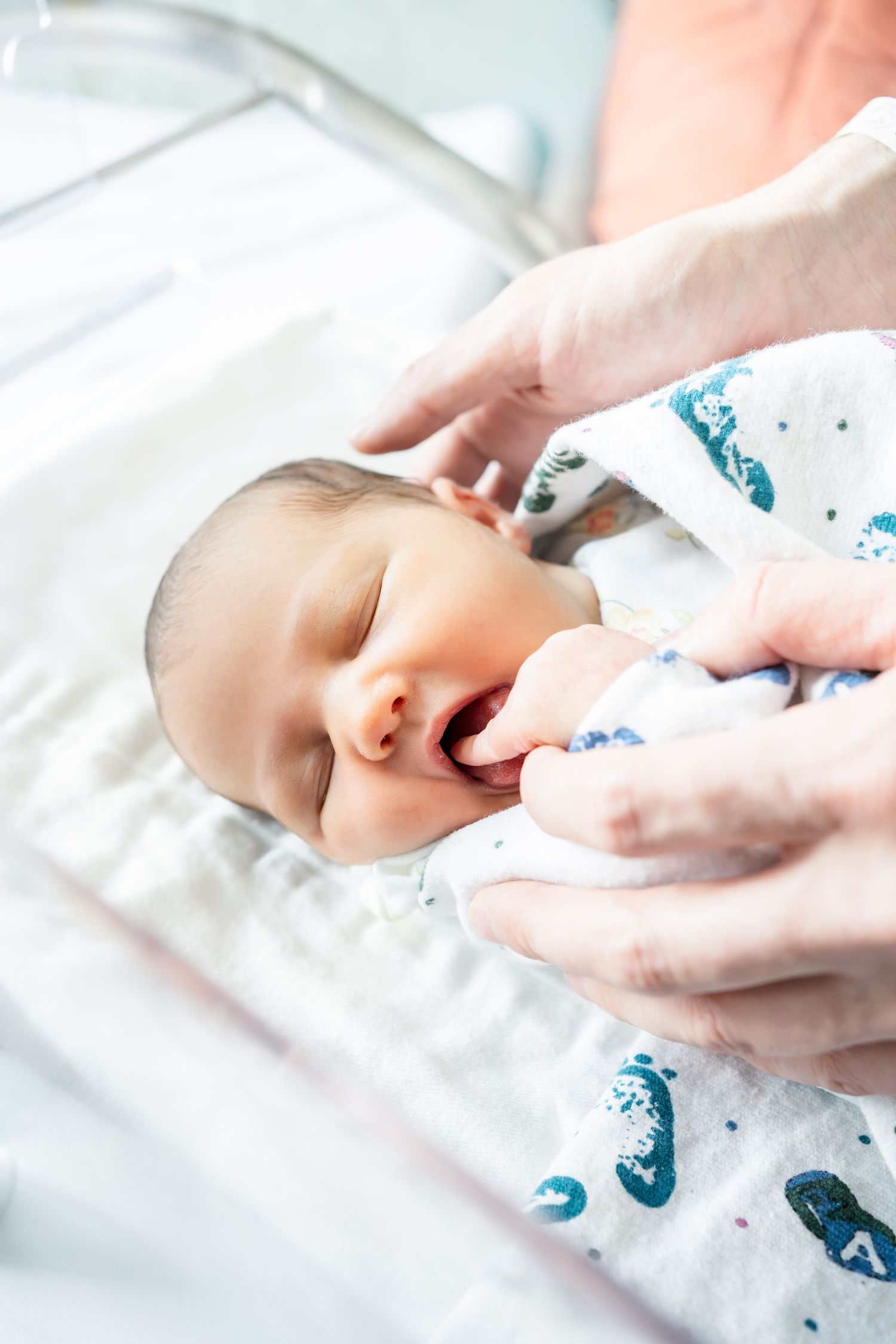 Newborn baby yawning while being held in a hospital bassinet, wrapped in a white patterned swaddle blanket.