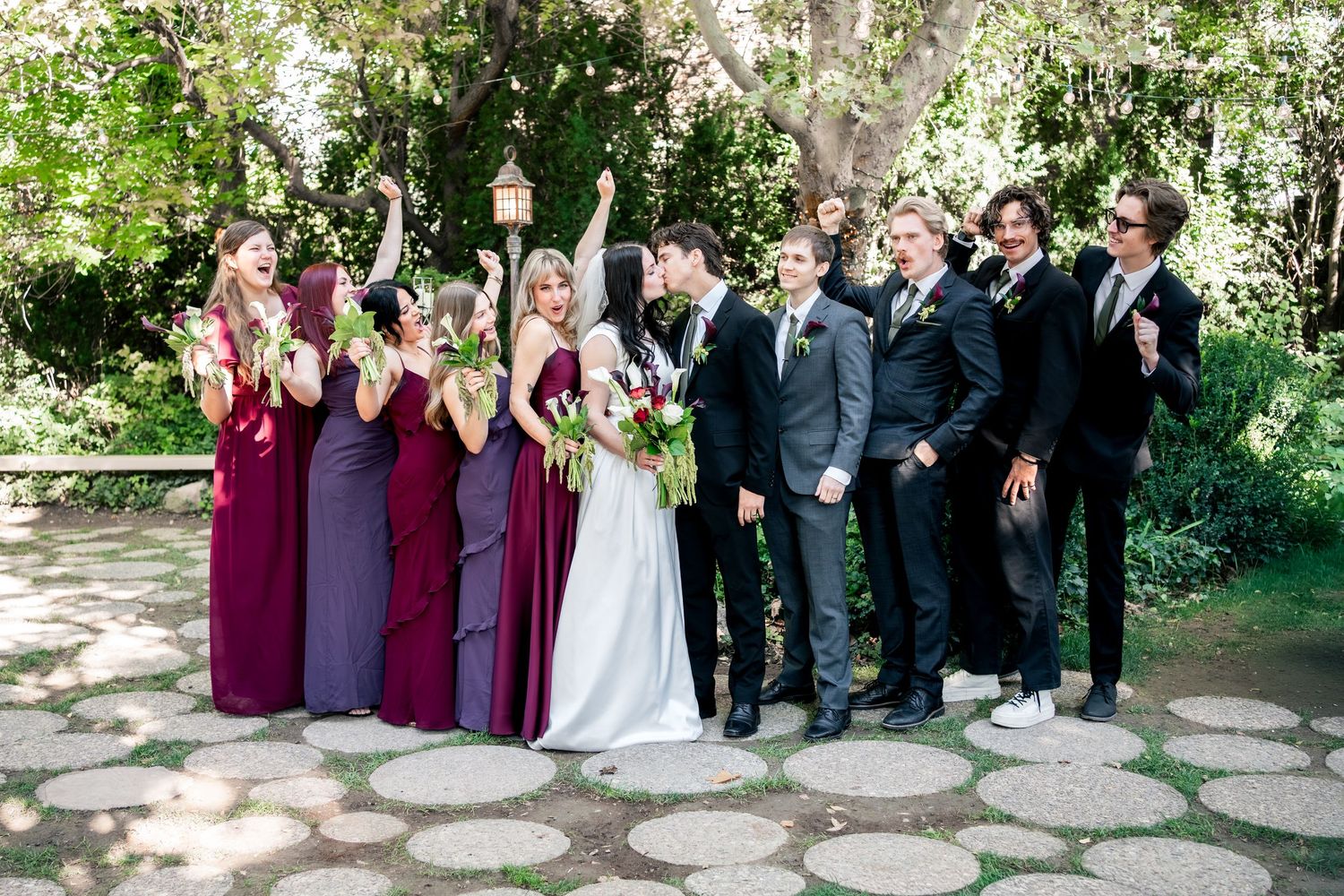 Bride in white gown with bridesmaids in burgundy and plum dresses and groomsmen in dark suits cheering on a stone path in a sunny garden.