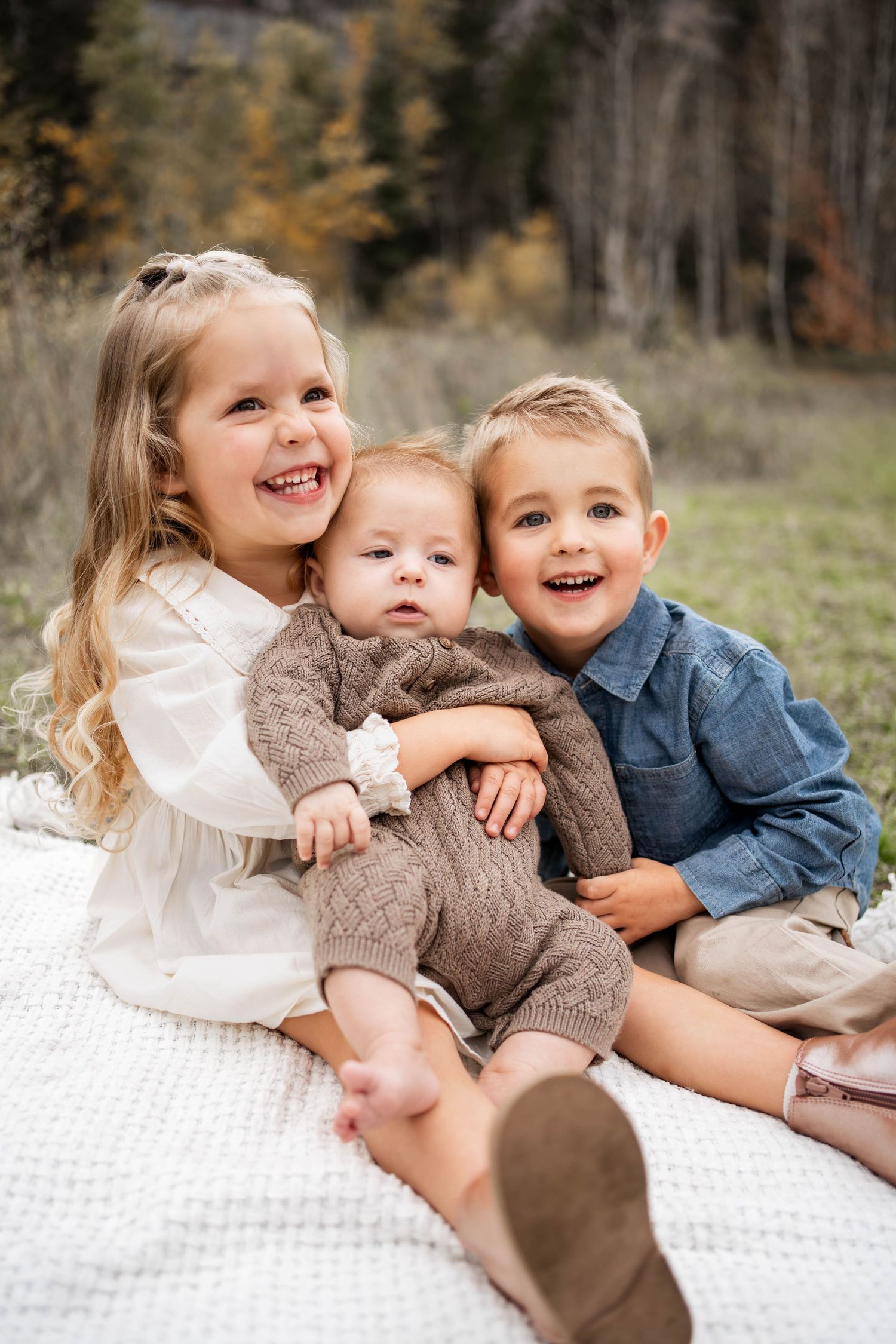 Three smiling young children cuddle on a white knit blanket outdoors, a girl holding a baby beside a boy in a denim shirt.