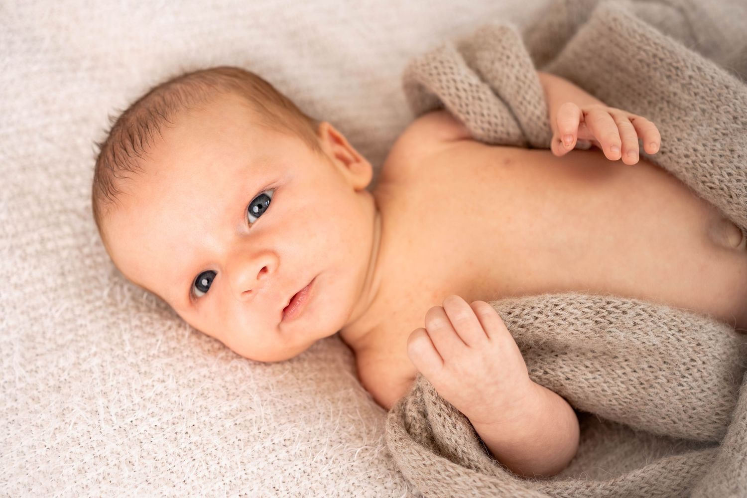 Newborn baby with bright eyes lying on a soft beige blanket, wrapped in a knitted taupe throw with tiny hands raised.