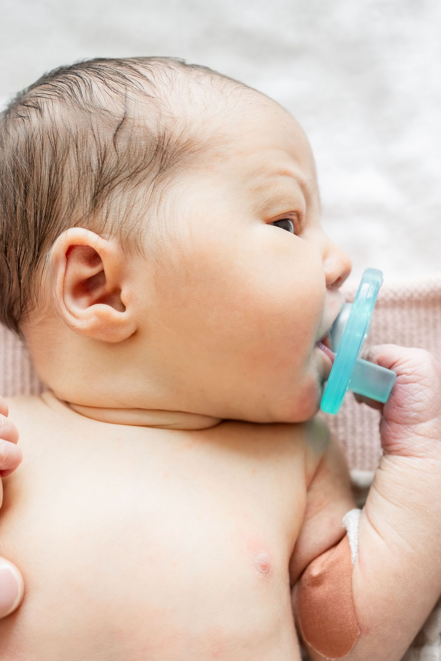Close-up of a newborn baby with dark hair sucking on a teal pacifier while lying on a soft blanket.
