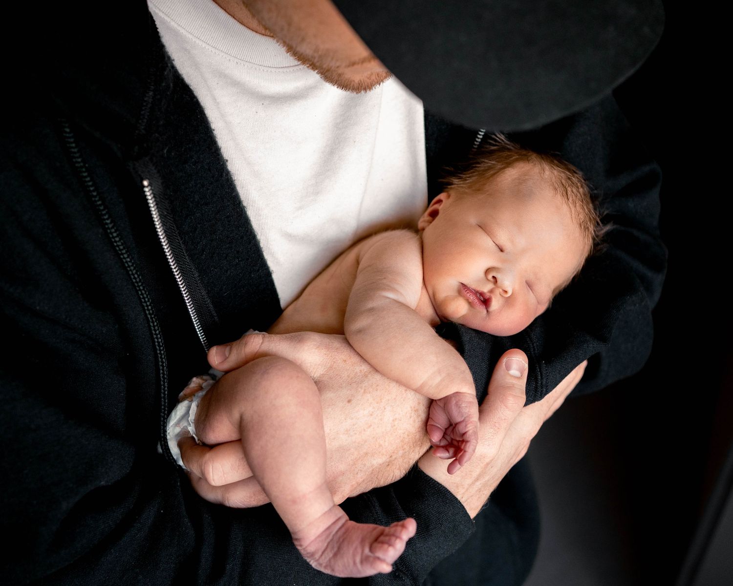 Newborn baby sleeping peacefully in parent's arms, cradled against a black jacket.