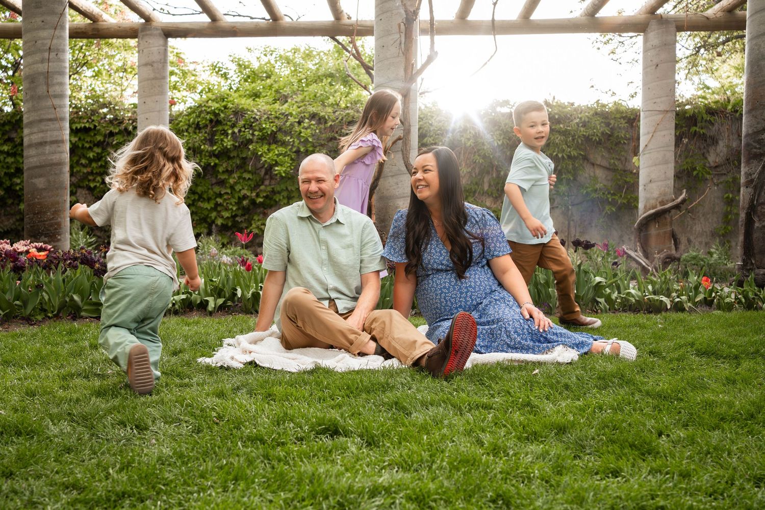 A joyful family sits on a blanket in a sunlit garden while three young children play around their smiling parents.