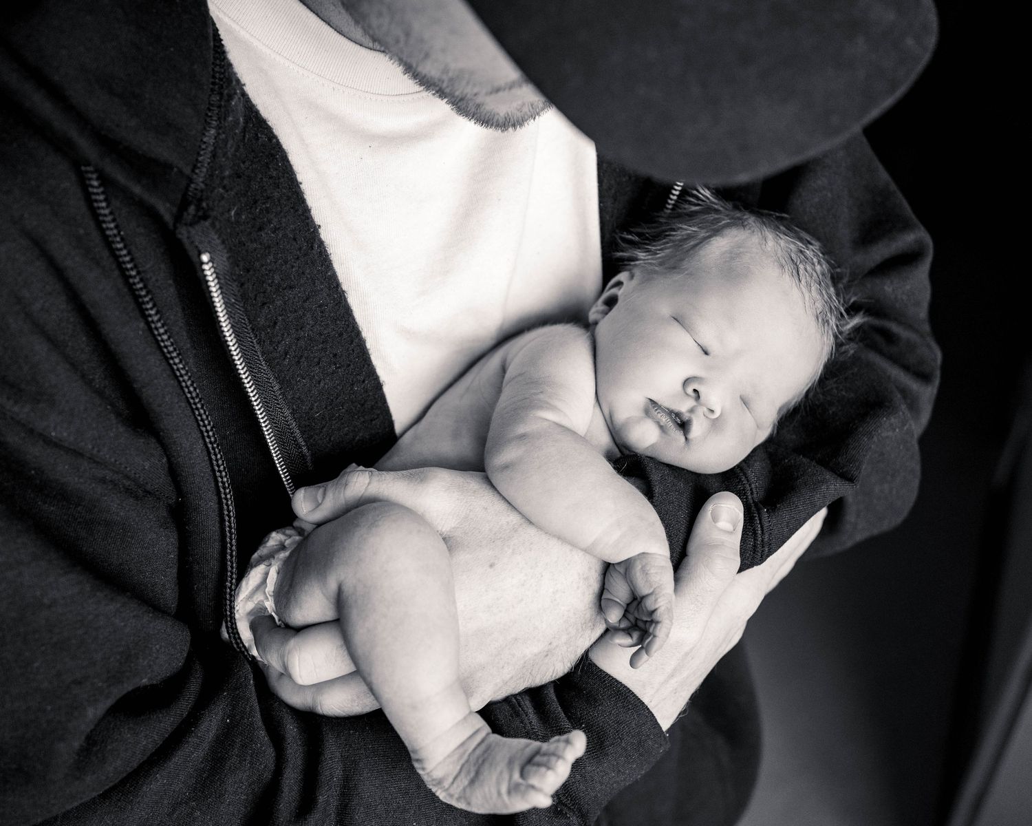 Black and white photo of a sleeping newborn baby cradled in adult hands against a dark jacket.